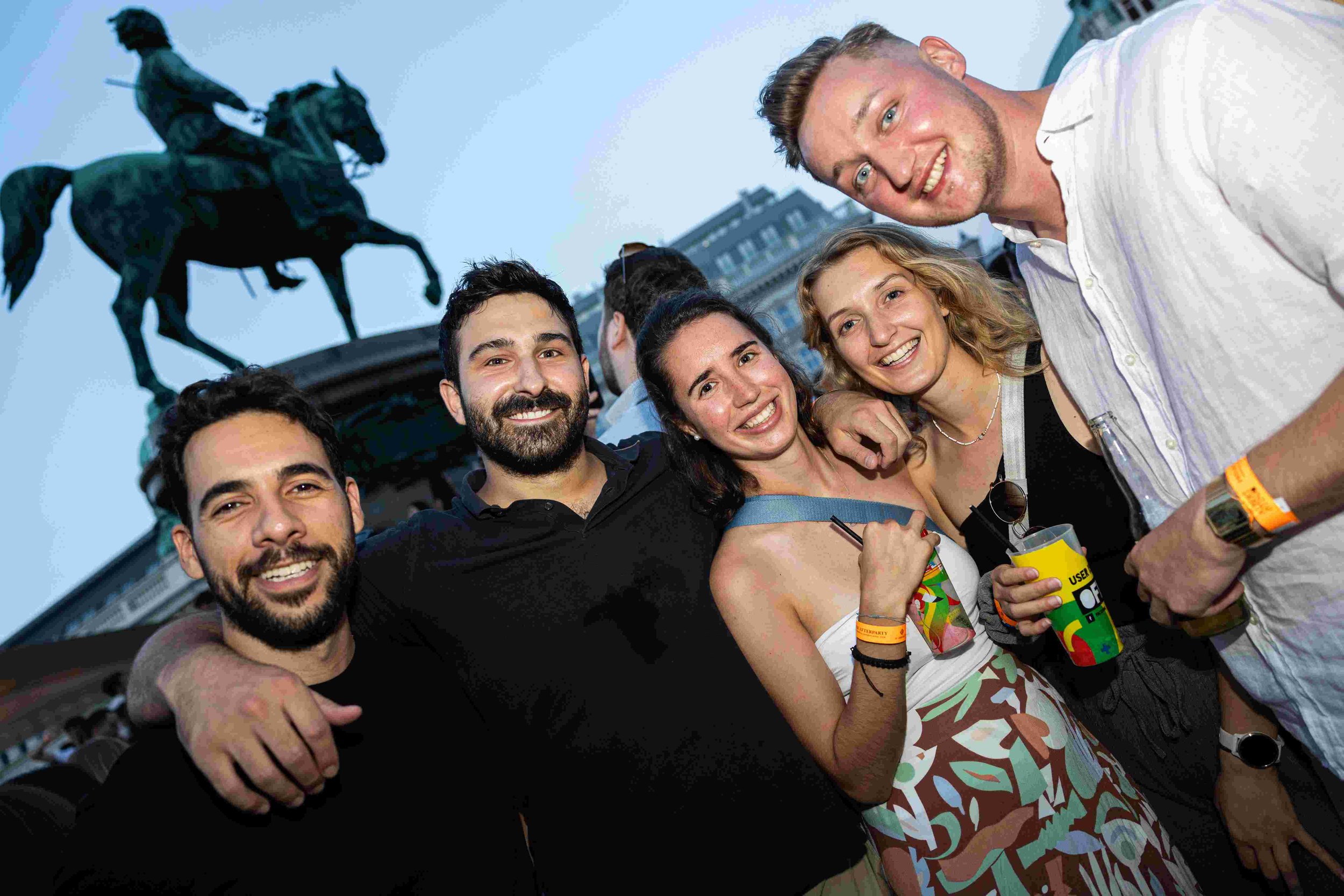 Group of six young adults taking a selfie outdoors with a cityscape and a statue of a horse and rider in the background, smiling and enjoying a social gathering.