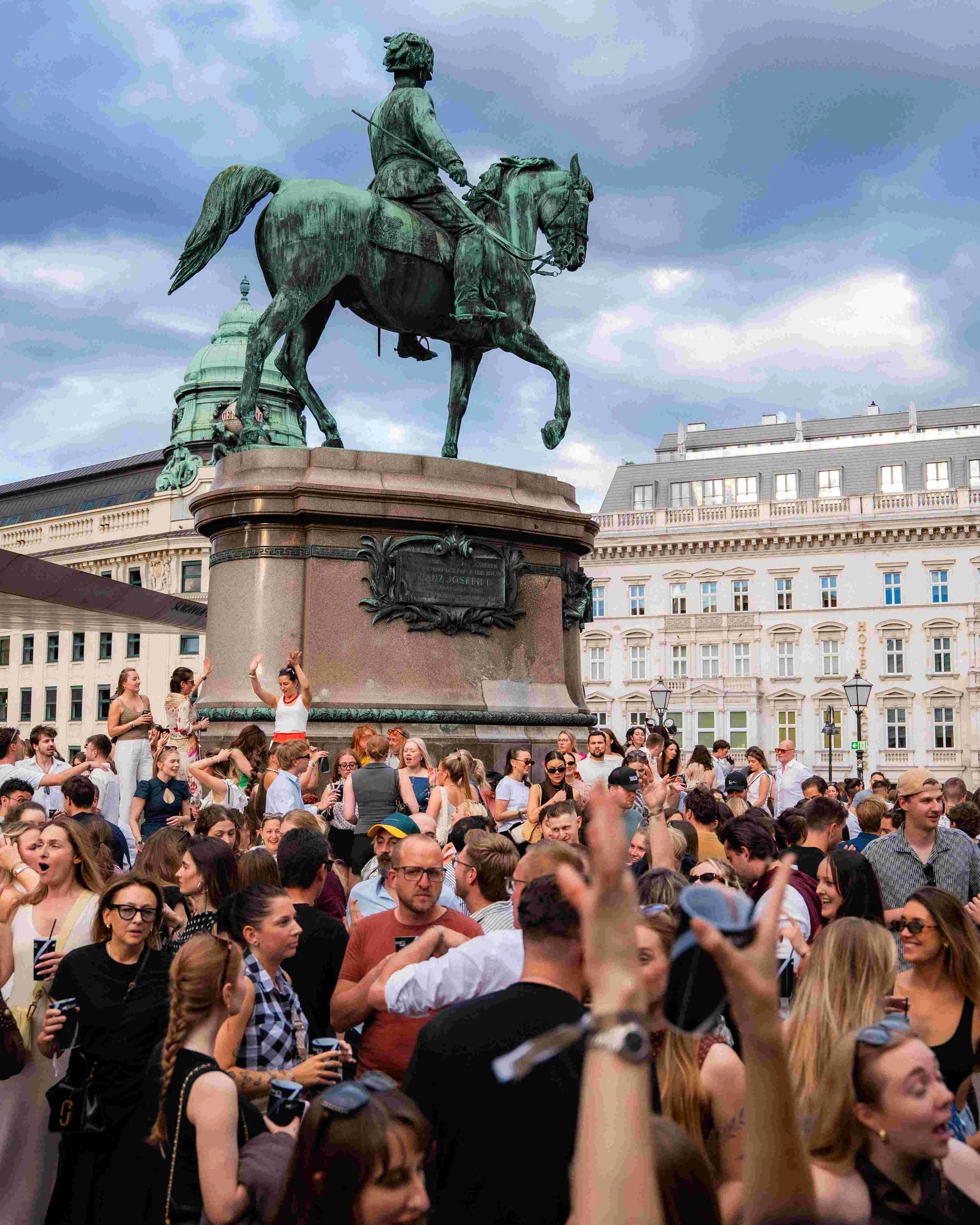 A crowded square filled with people surrounding a large bronze equestrian statue of a man in historical clothing, with buildings and cloudy sky in the background.