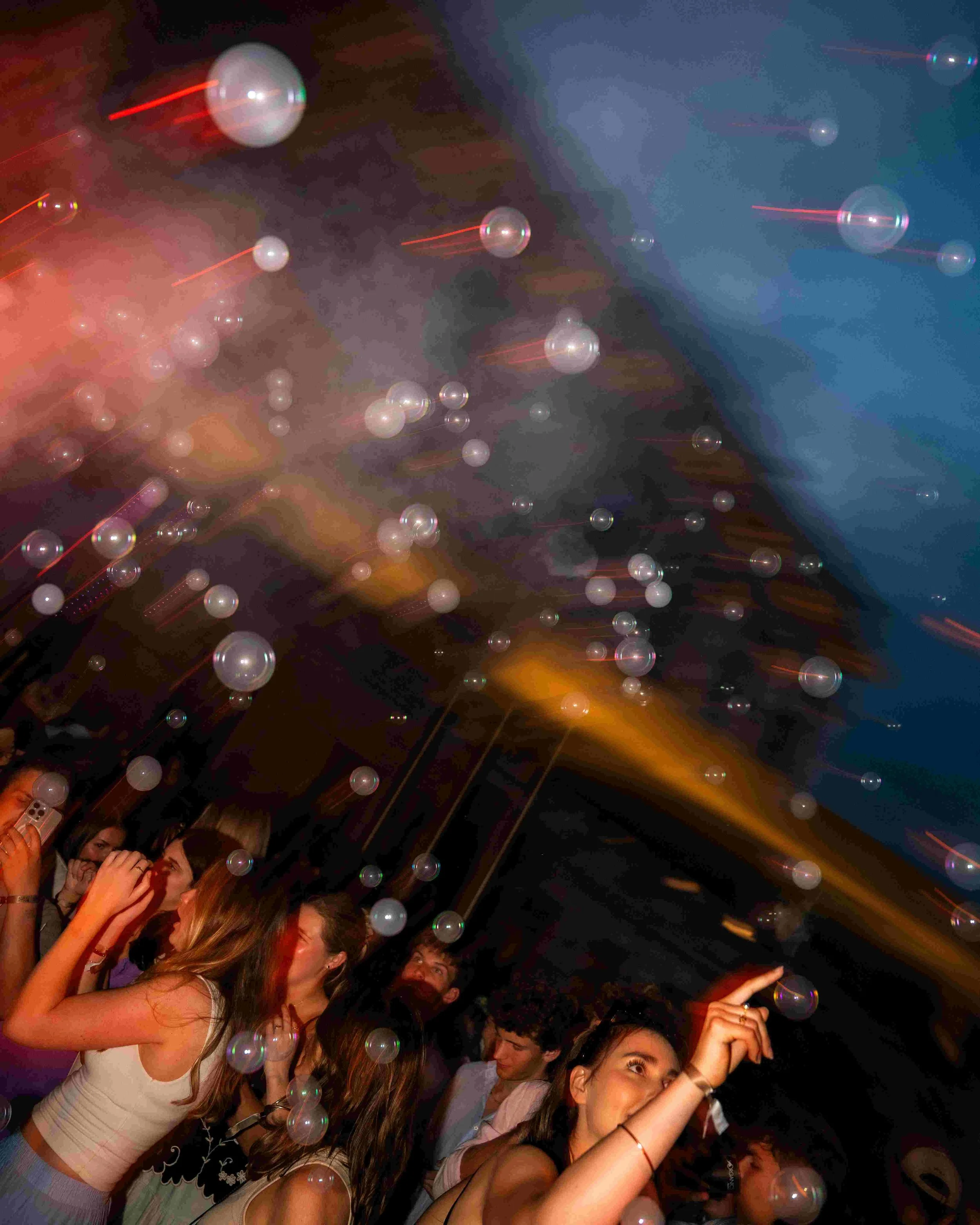 A crowd of young people dancing and celebrating at a party or nightclub, with bubbles floating in the air and colorful lighting creating a lively atmosphere.