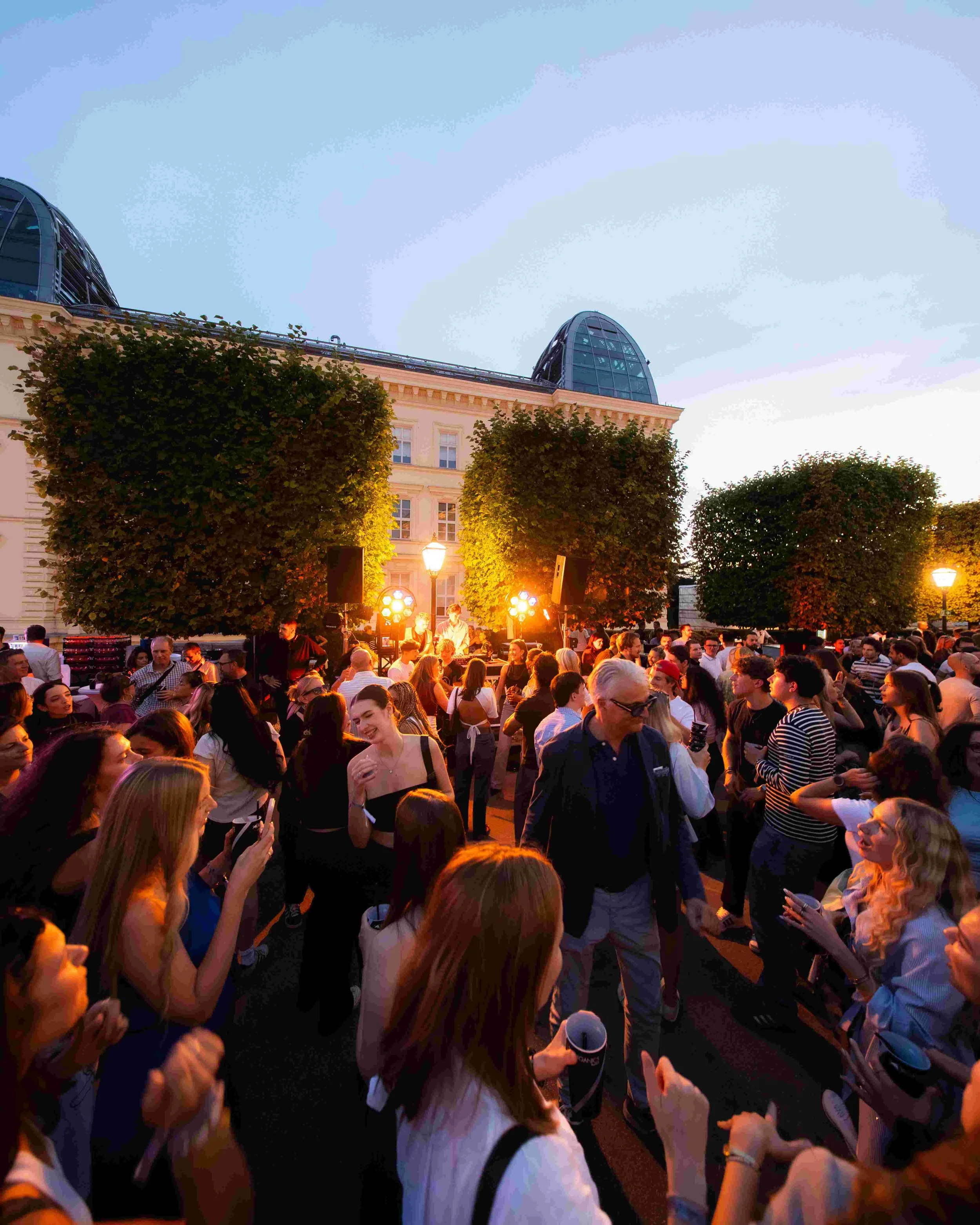 A large outdoor crowd at dusk with people socializing and enjoying a gathering, illuminated by streetlights and surrounded by trees and classic buildings.