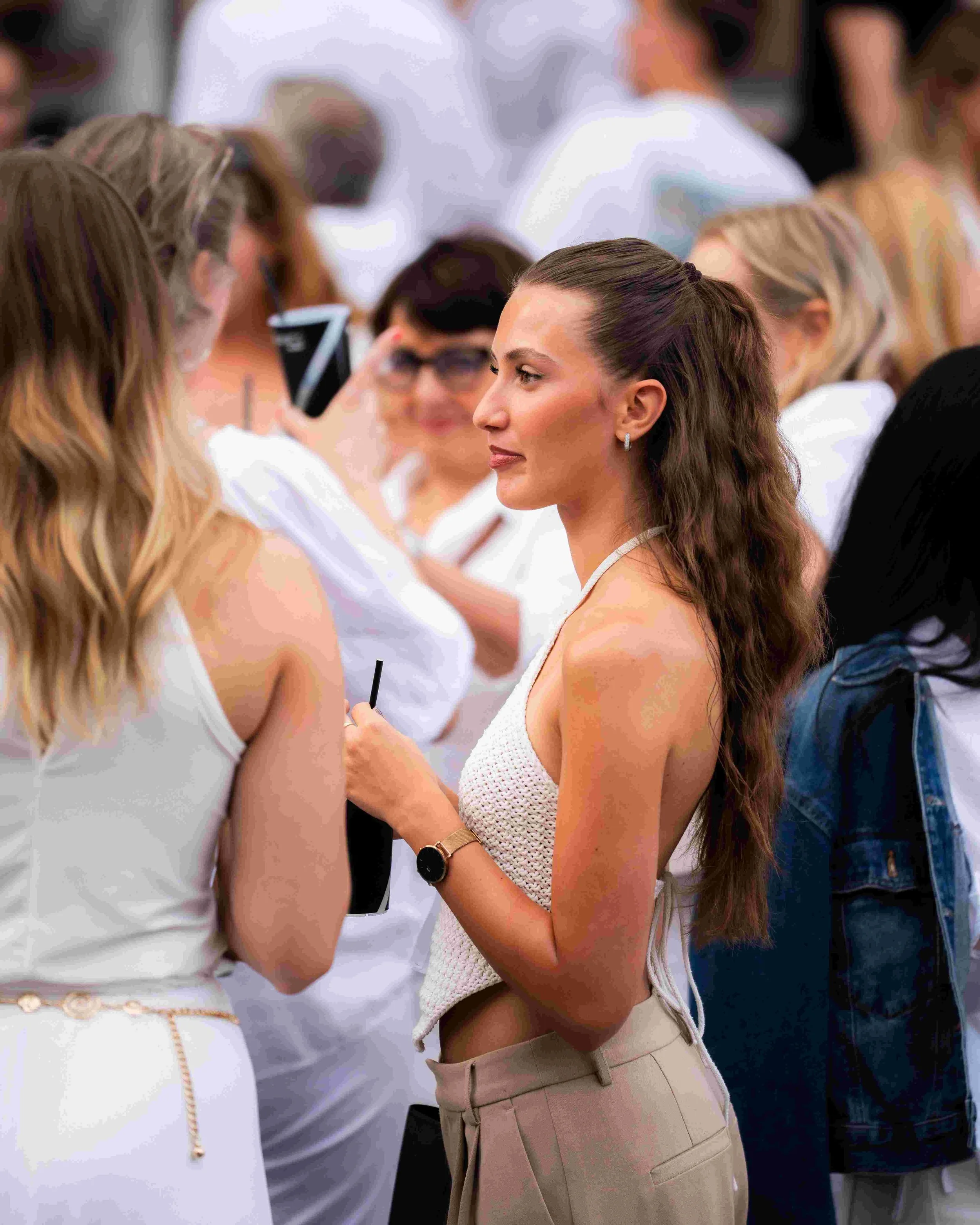 A group of women engaging in conversation at a social event, with focus on a brunette woman with long wavy hair. She is wearing a white textured sleeveless top, beige high-waisted trousers, a gold watch, and small hoop earrings.