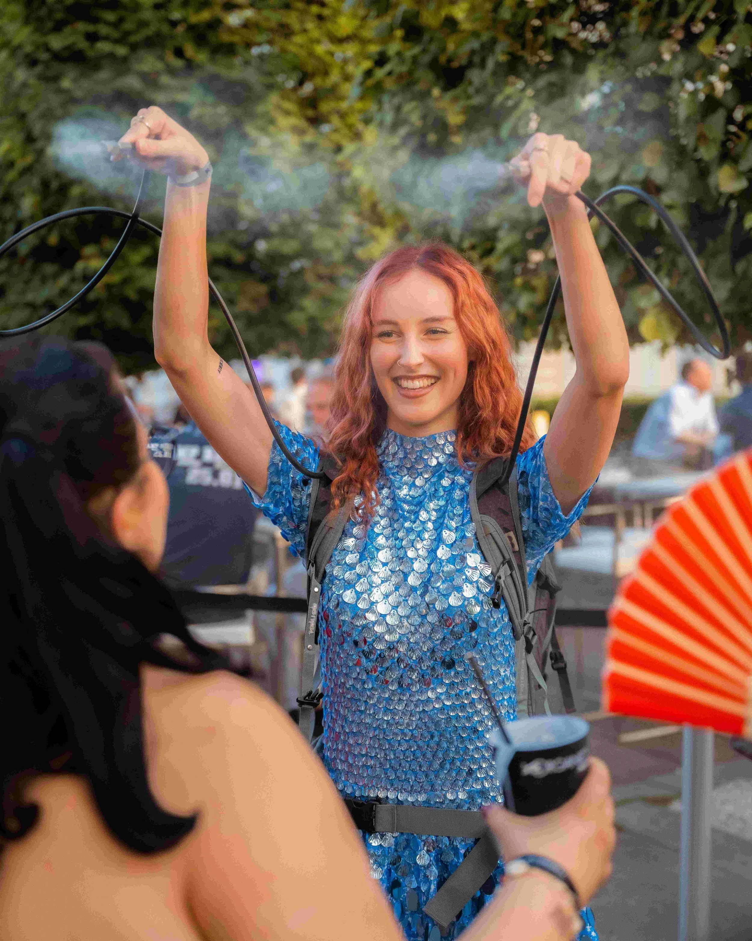 A woman with red curly hair smiling and raising her arms, holding a vape device emitting vapor, wearing a blue shiny dress, with guests in the background at an outdoor event.