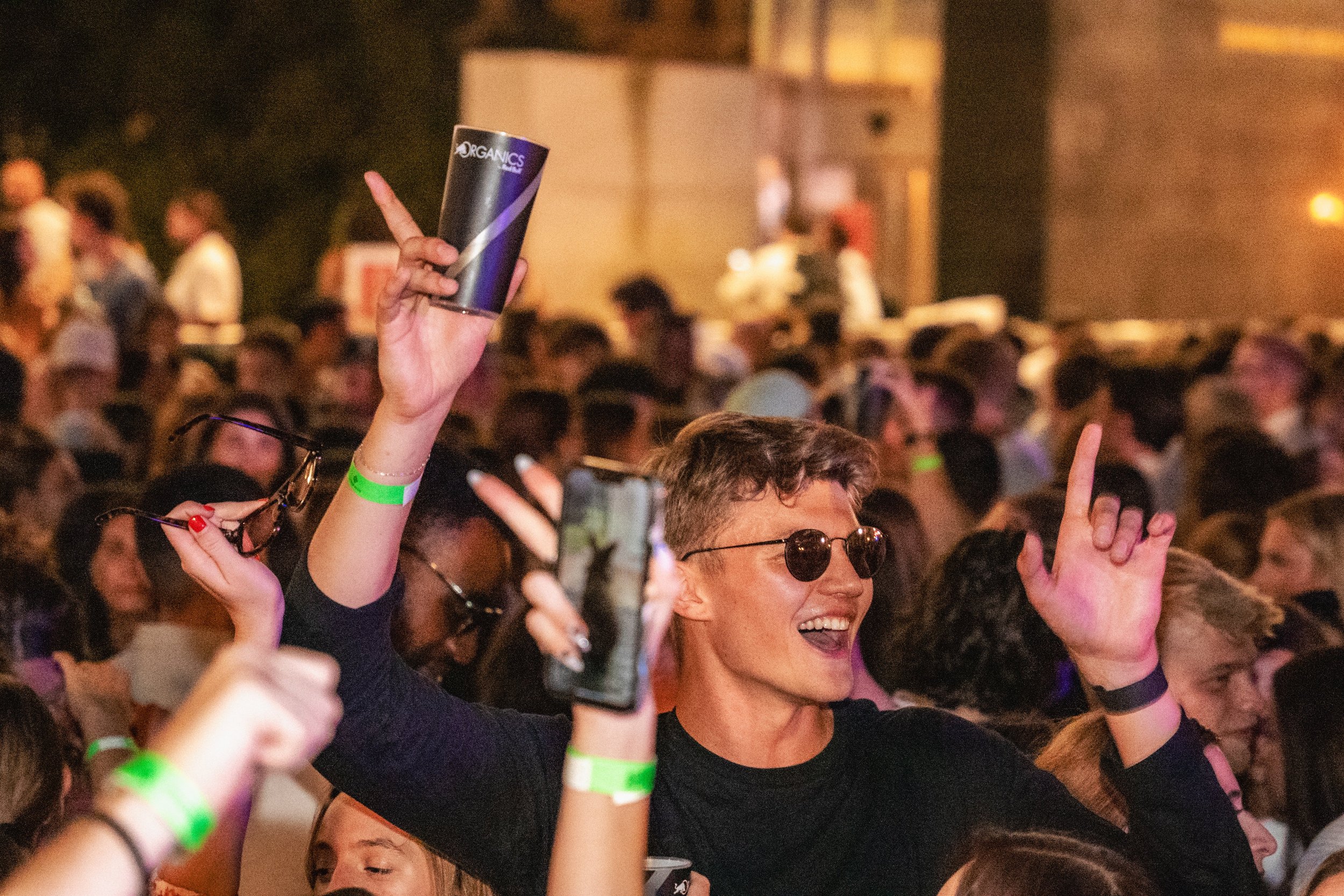 Crowd of people at a lively event, some raising their hands, with one person holding a black cup labeled 'ORGANICS'. Many wearing green wristbands.