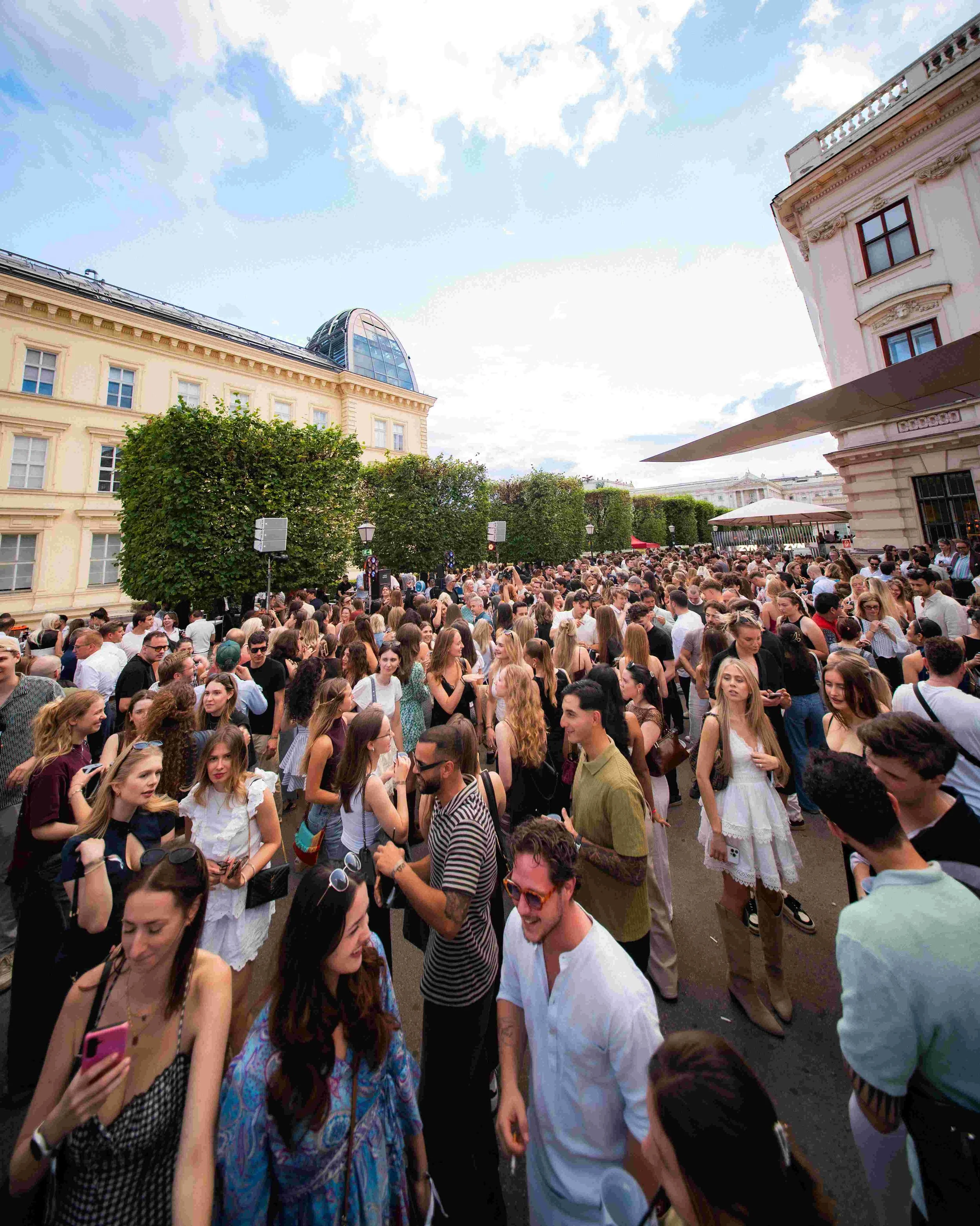 A large crowd of people gathered outdoors in a city area with historic buildings and a large glass dome structure in the background. The sky is partly cloudy.