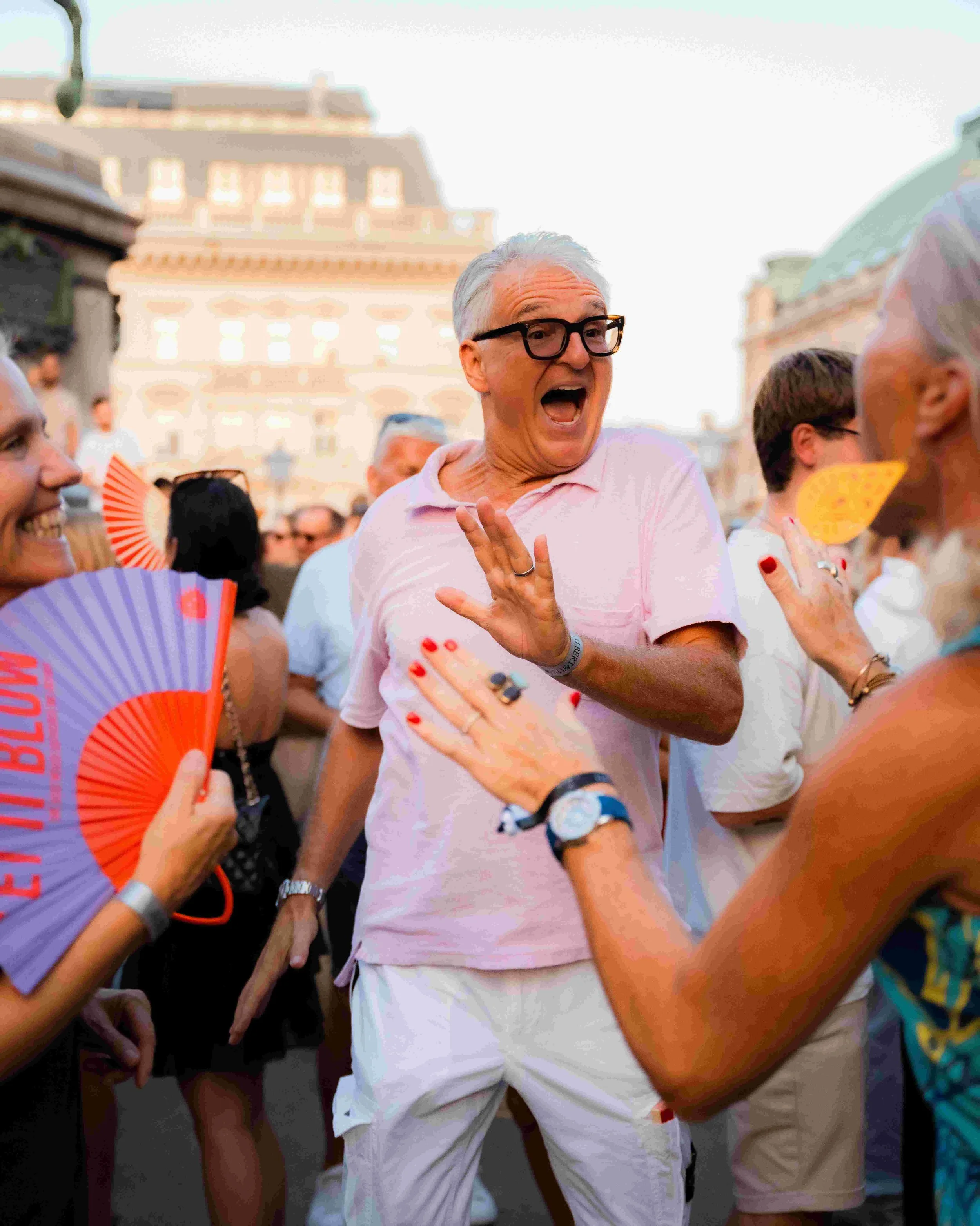 An older man with gray hair and black glasses, wearing a pink shirt and white shorts, appears to be yelling or shouting during a crowded outdoor event. He is surrounded by other people, some holding colorful fans, and the background shows historic ar