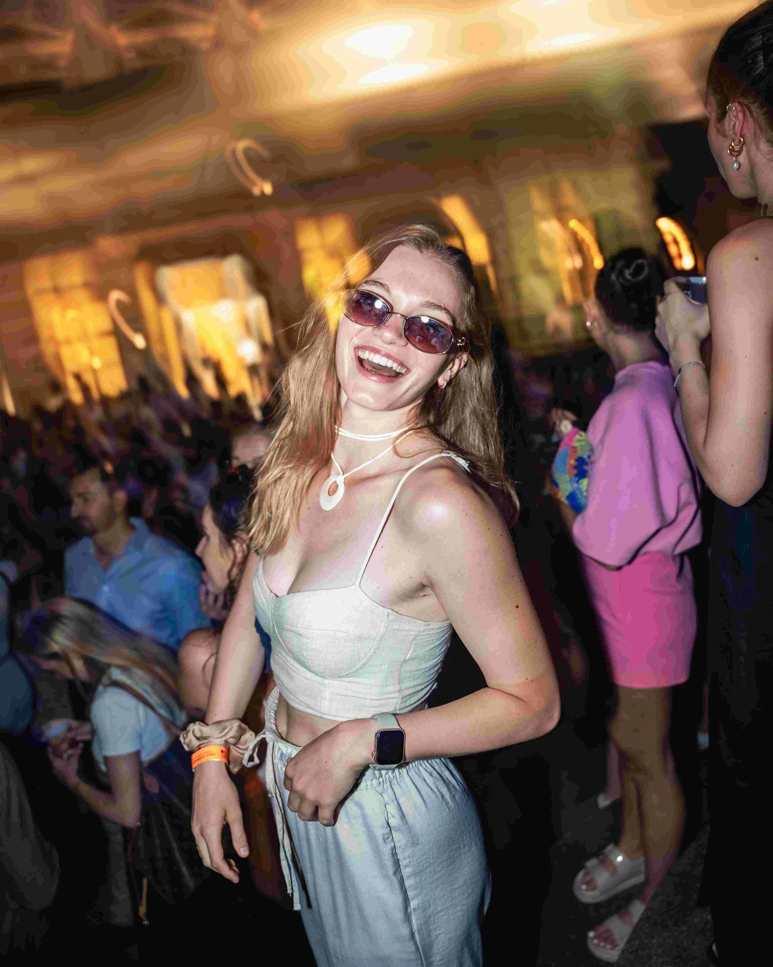 A young woman with long blonde hair wearing sunglasses, a white spaghetti strap top, and light blue striped pants is smiling and dancing at a party or nightclub. She is wearing an Apple Watch and a white choker necklace. The background shows other wo