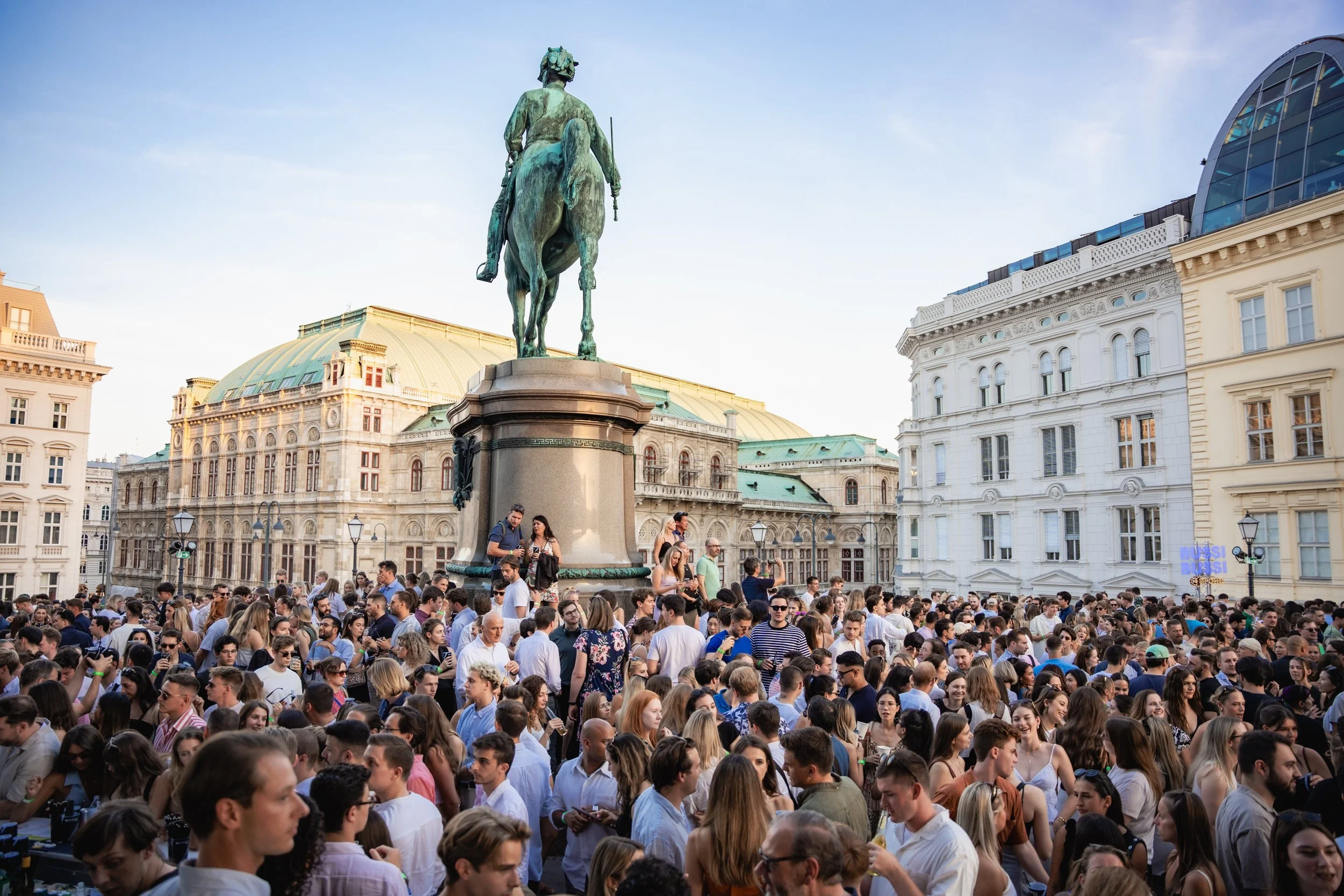 Crowd gathered around a statue of a man on a horse in an urban square, with historic buildings and a modern glass roof structure in the background.