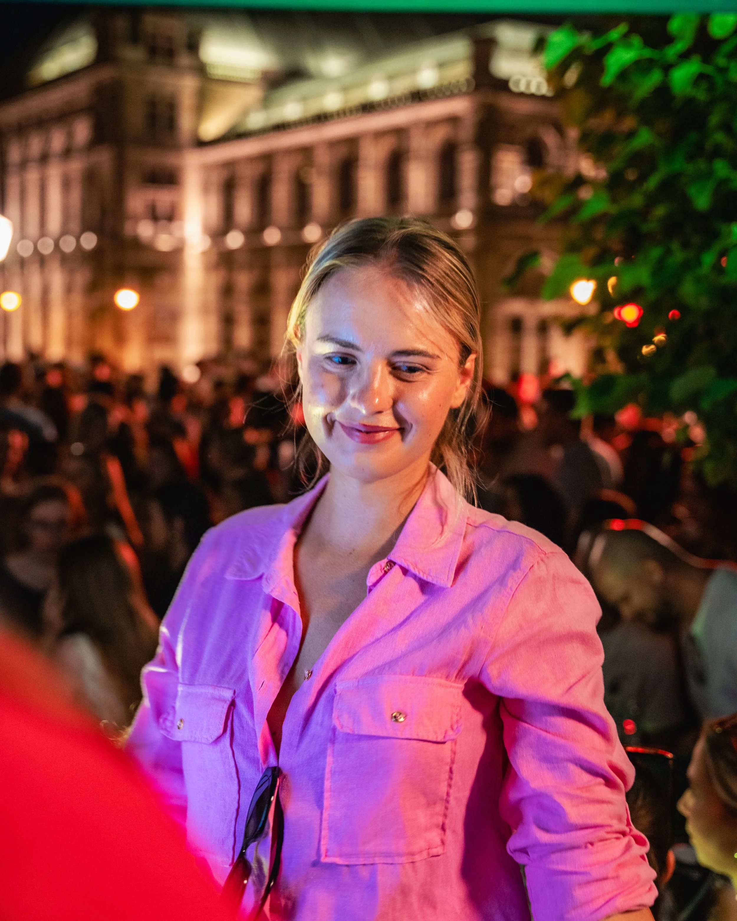 A young woman smiling at night in an outdoor setting with a large historic building illuminated in the background, surrounded by greenery and city lights.