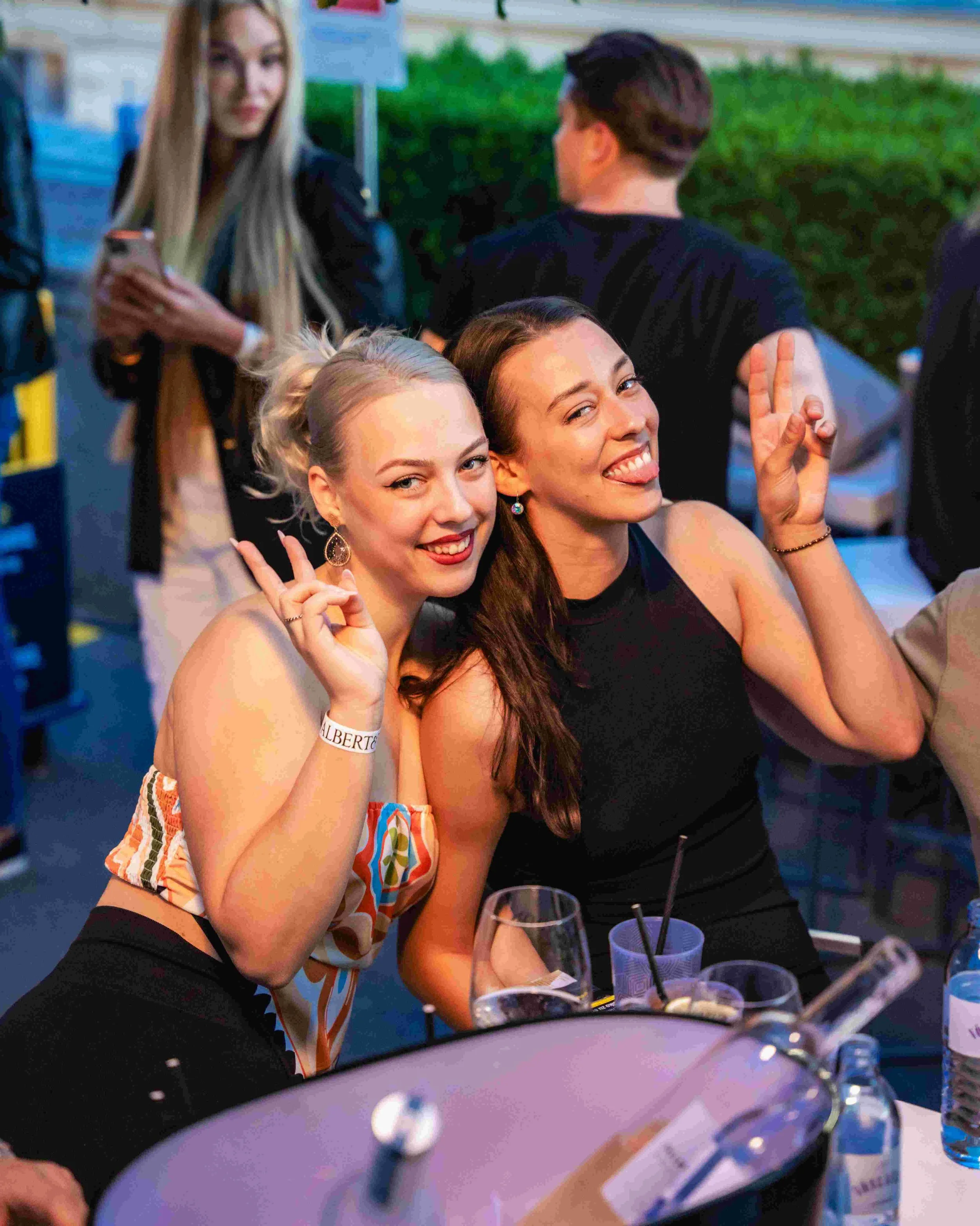 Two women at a social gathering, both smiling and making peace signs with their hands, with drinks on the table in front of them. The background includes other people and greenery.