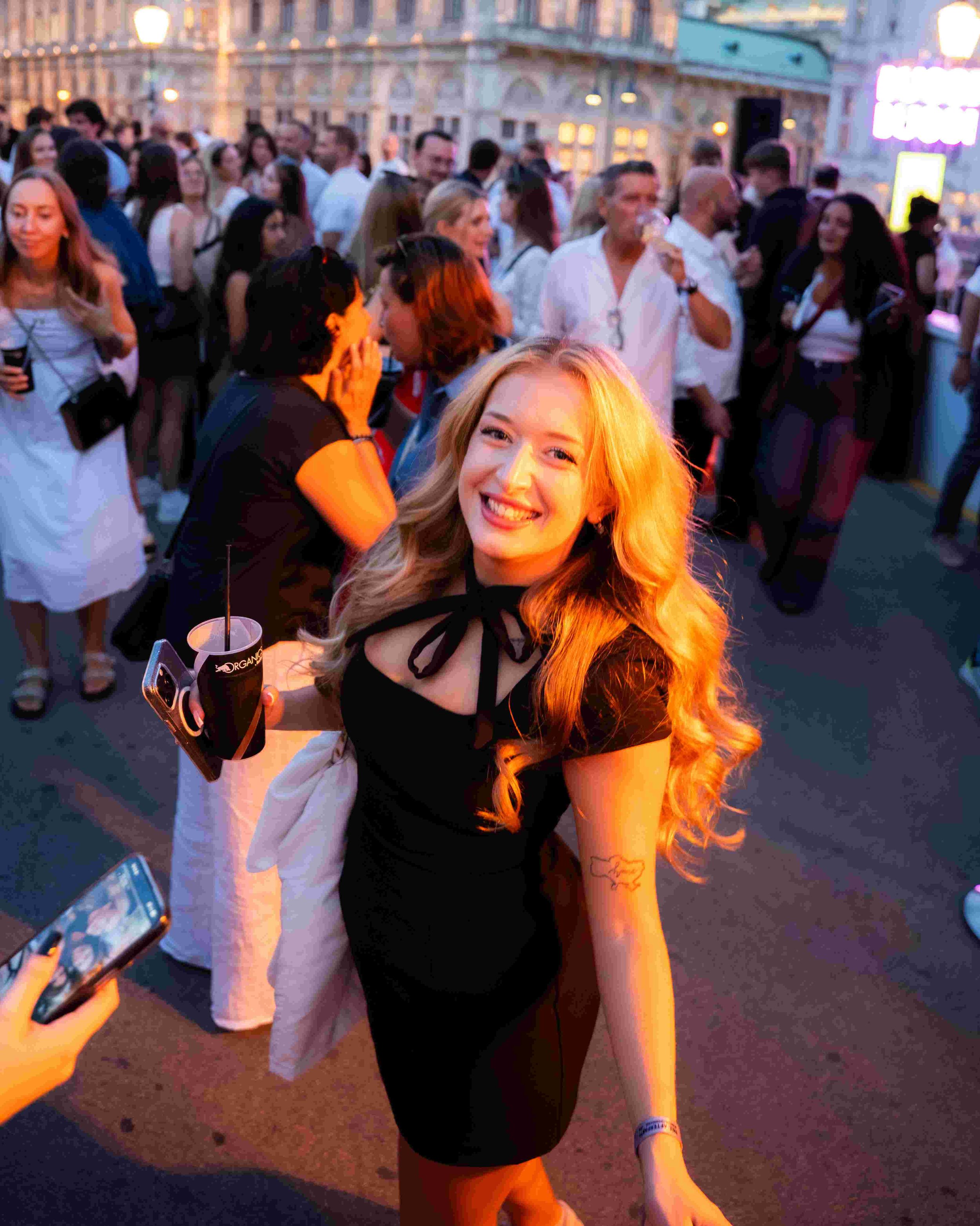 A smiling young woman with long blond hair wearing a black dress with sheer sleeves is at an outdoor event crowded with people. She is holding a drink in her left hand and a cellphone in her right hand. The background shows many people socializing ne