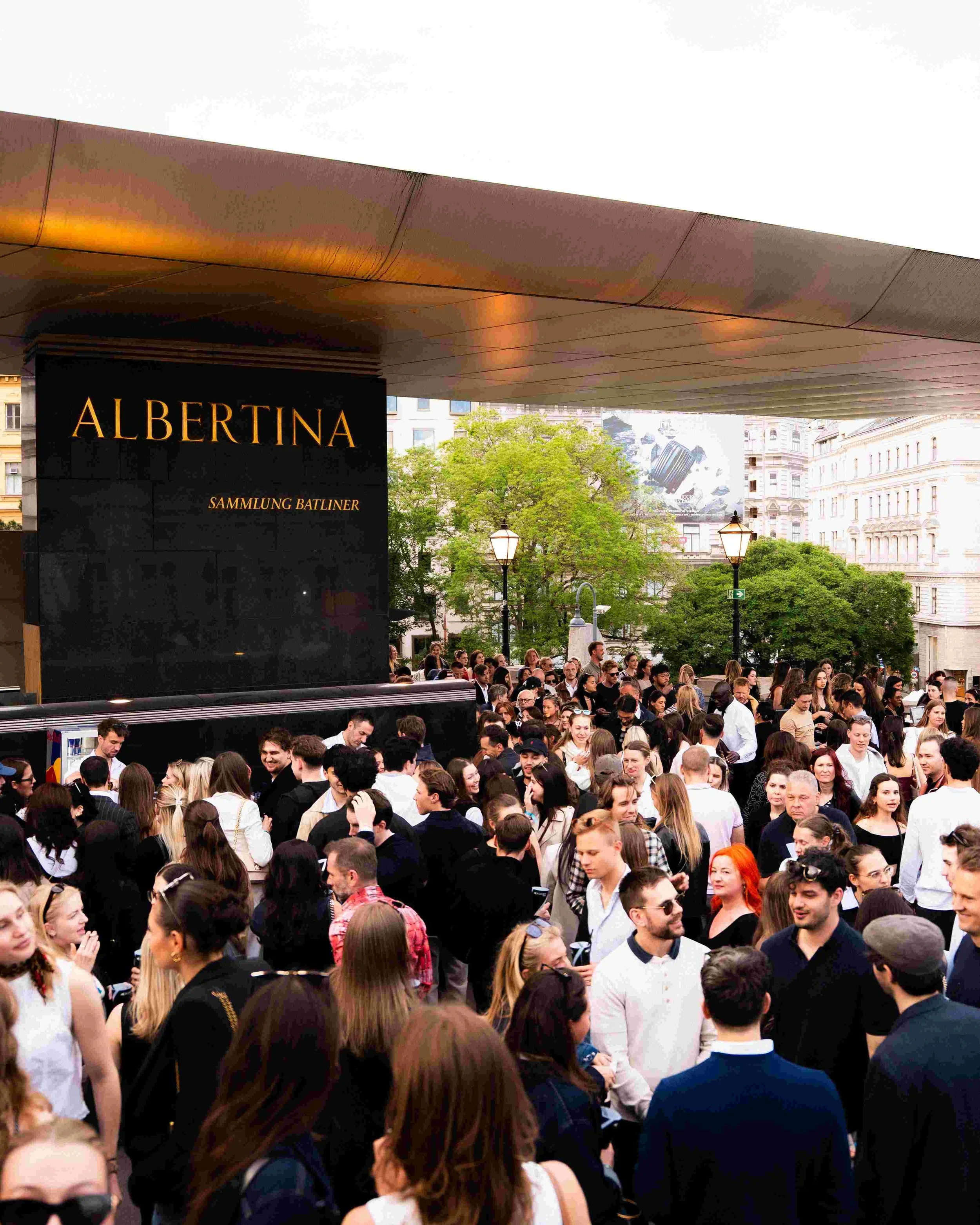 A crowd of people gathered outside beneath a large overhang, with a black sign that reads 'ALBERTINA SAMMLUNG BATLINER' and a cityscape with trees and buildings in the background.