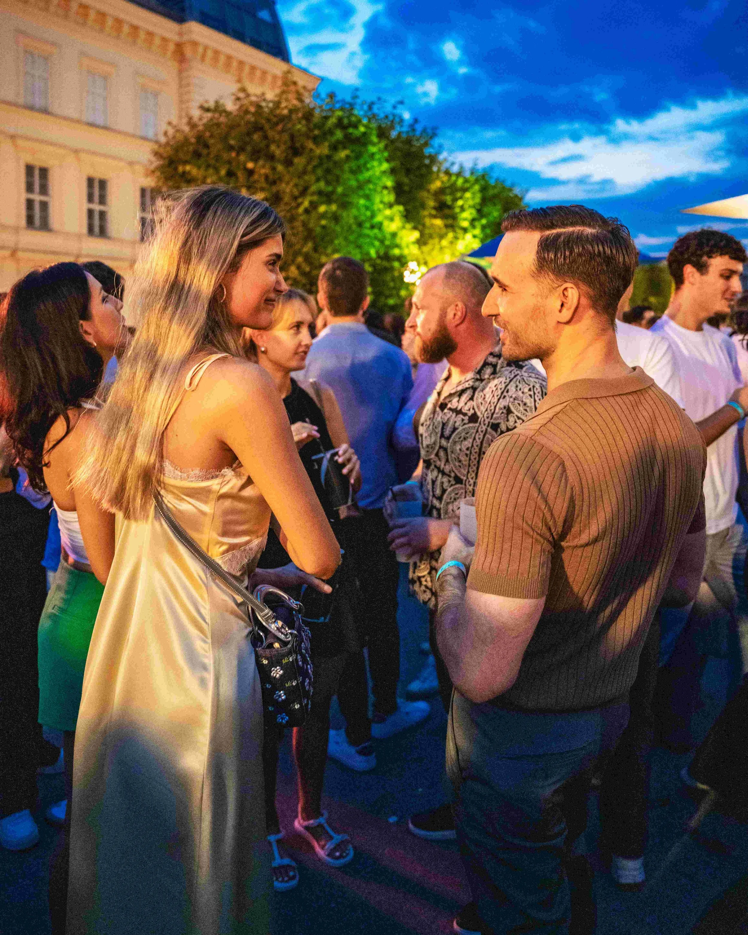People gathered outdoors in the evening, socializing and talking with each other, with a building and trees in the background.