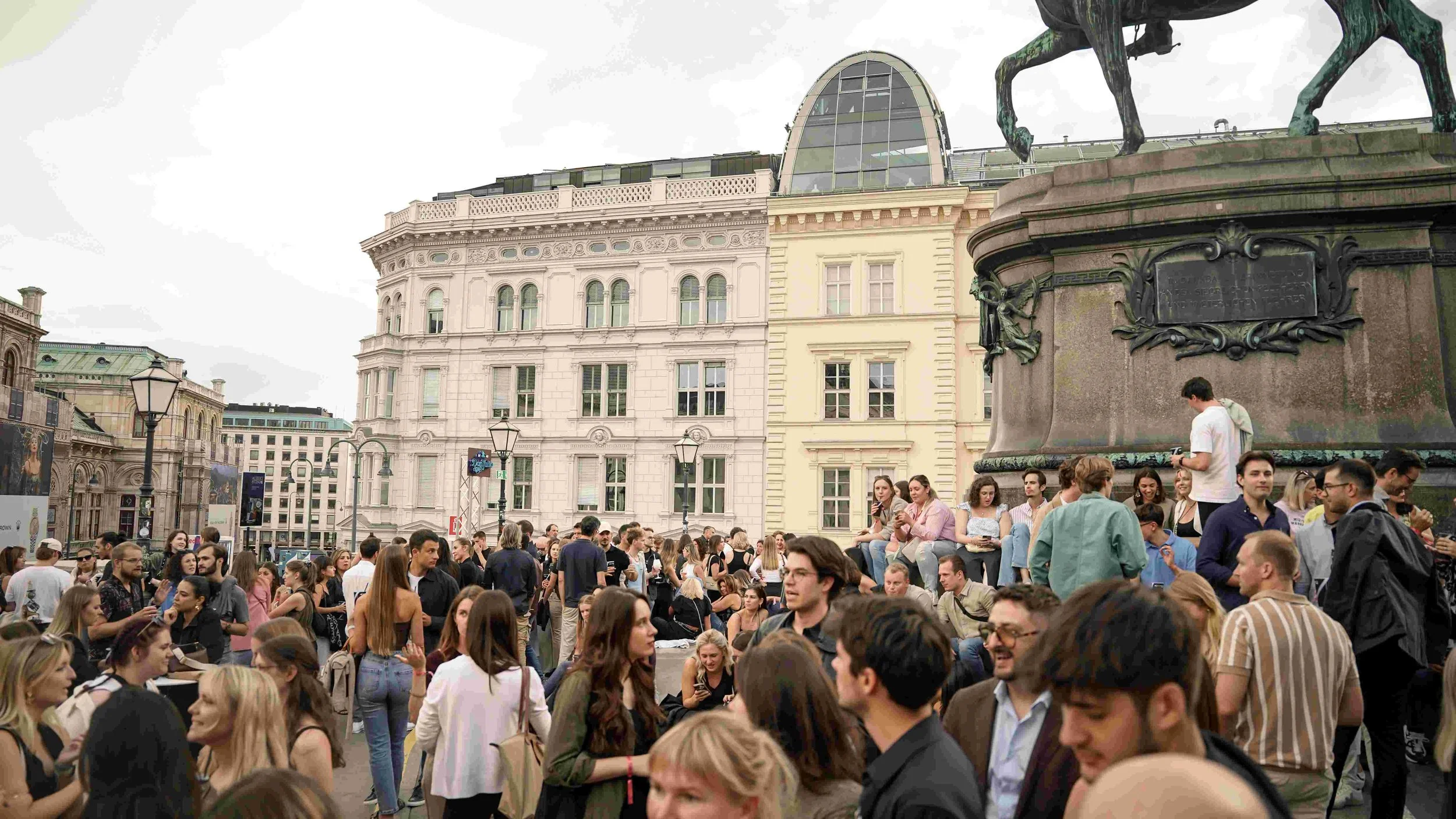 Crowd of people gathering in a city square with historic buildings in the background and a large bronze statue of a horse on a pedestal.