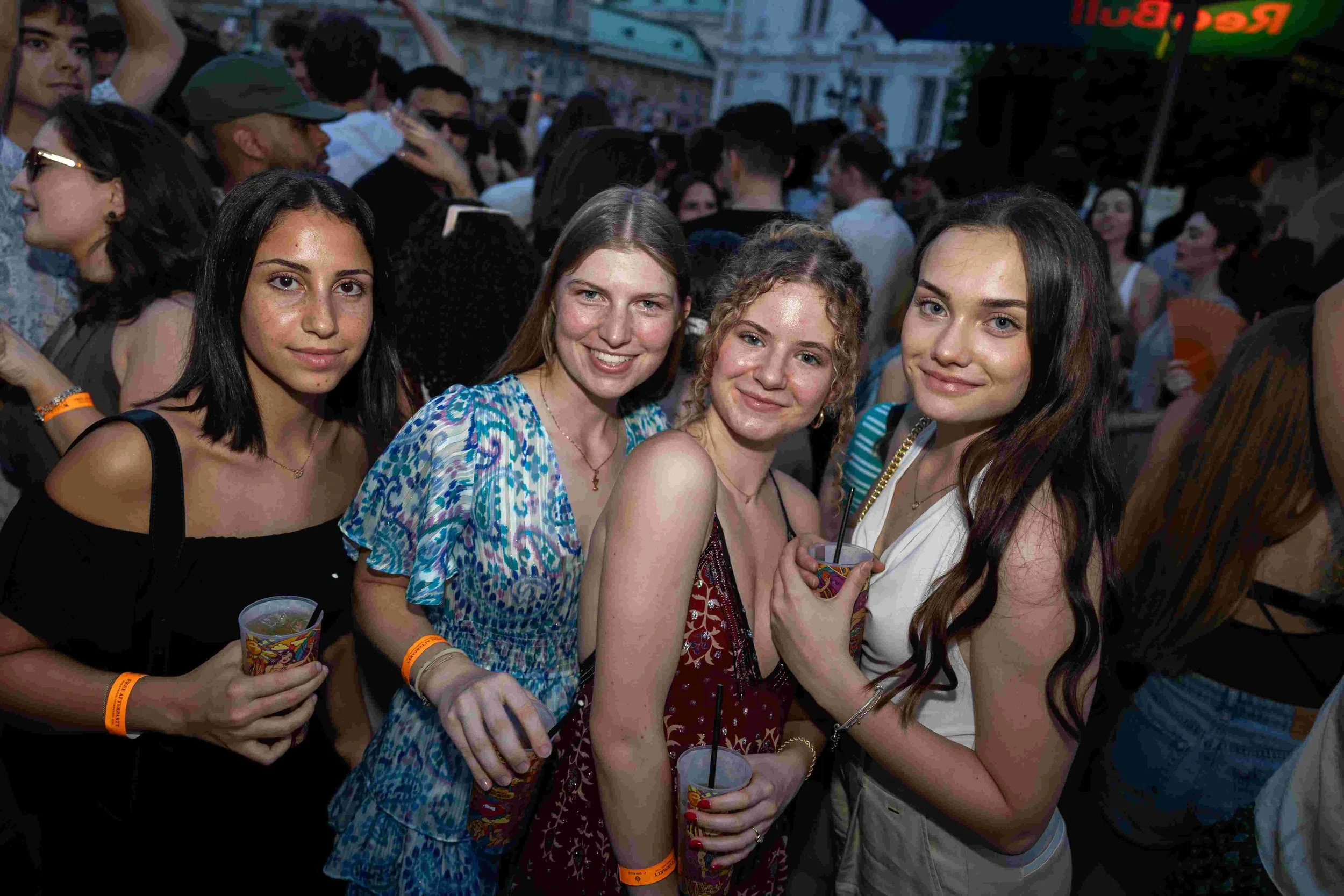 Four young women smiling at a crowded outdoor event, holding drinks in colorful cups, wearing casual summer clothes, with many people behind them.
