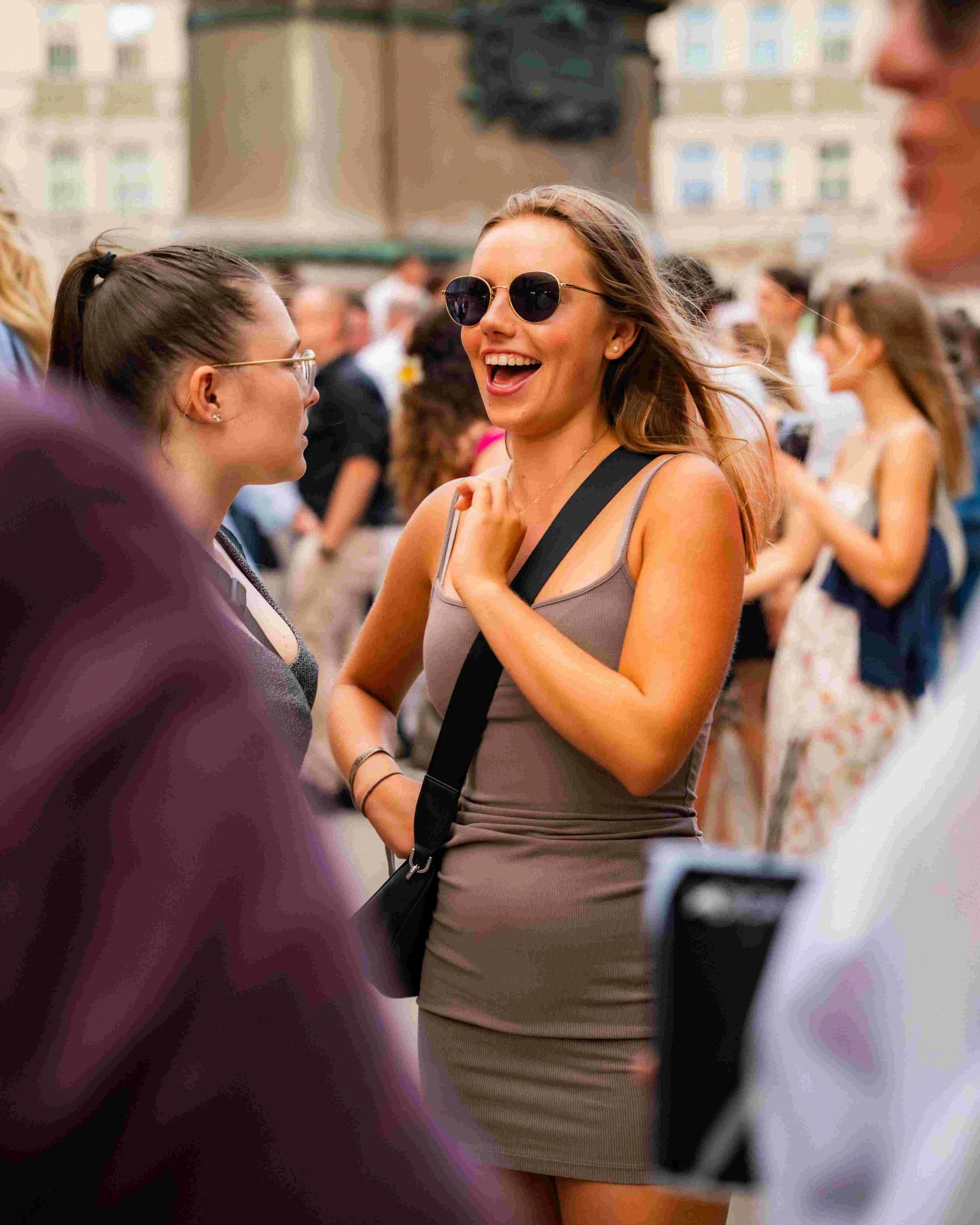 A woman wearing sunglasses and a beige dress, smiling, in a crowded outdoor event.