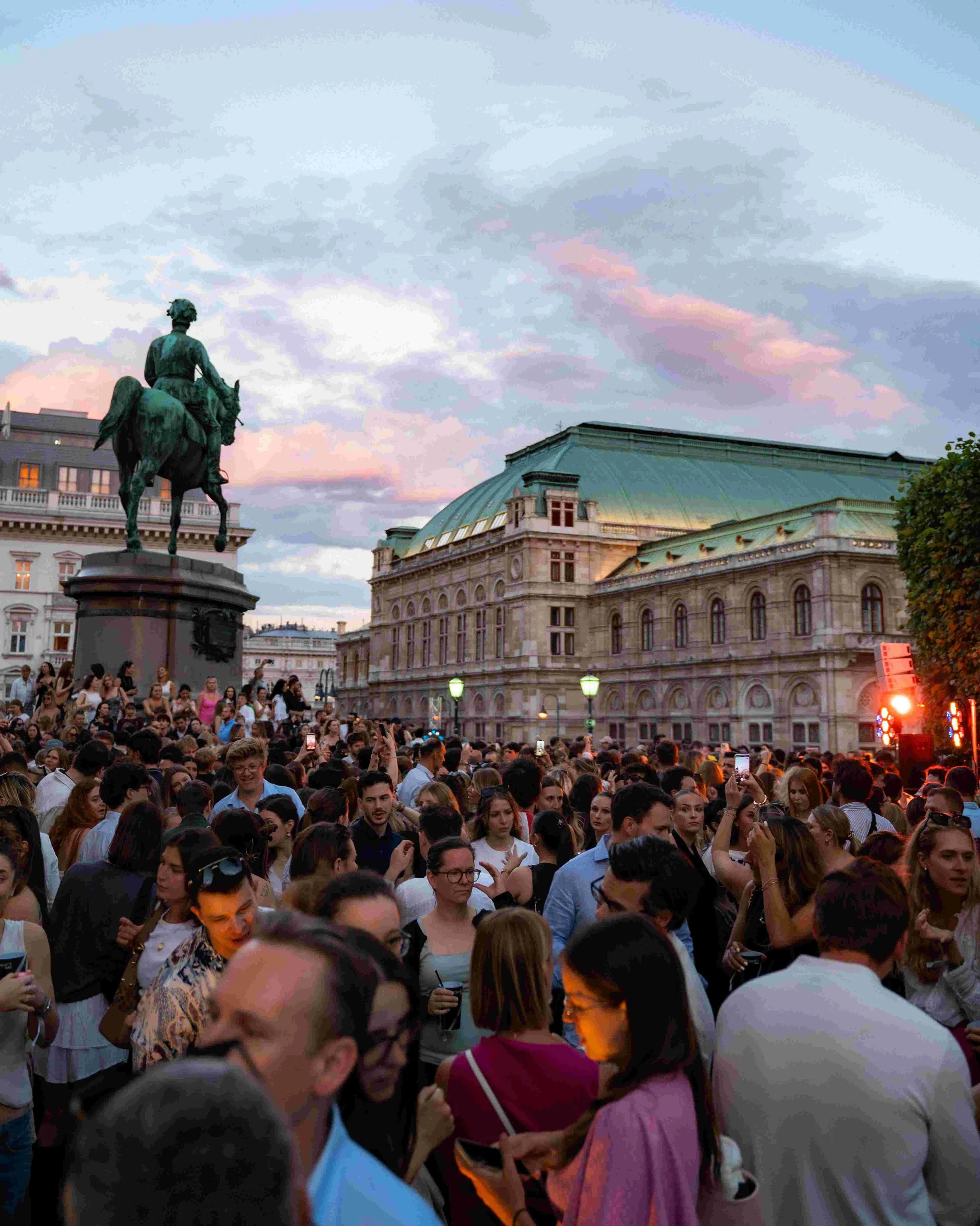 Large crowd of people gathered in an outdoor square, with a statue of a man on horseback on a pedestal in the background, under a sky with pink and blue clouds, and a historic building with a green roof and ornate architecture.