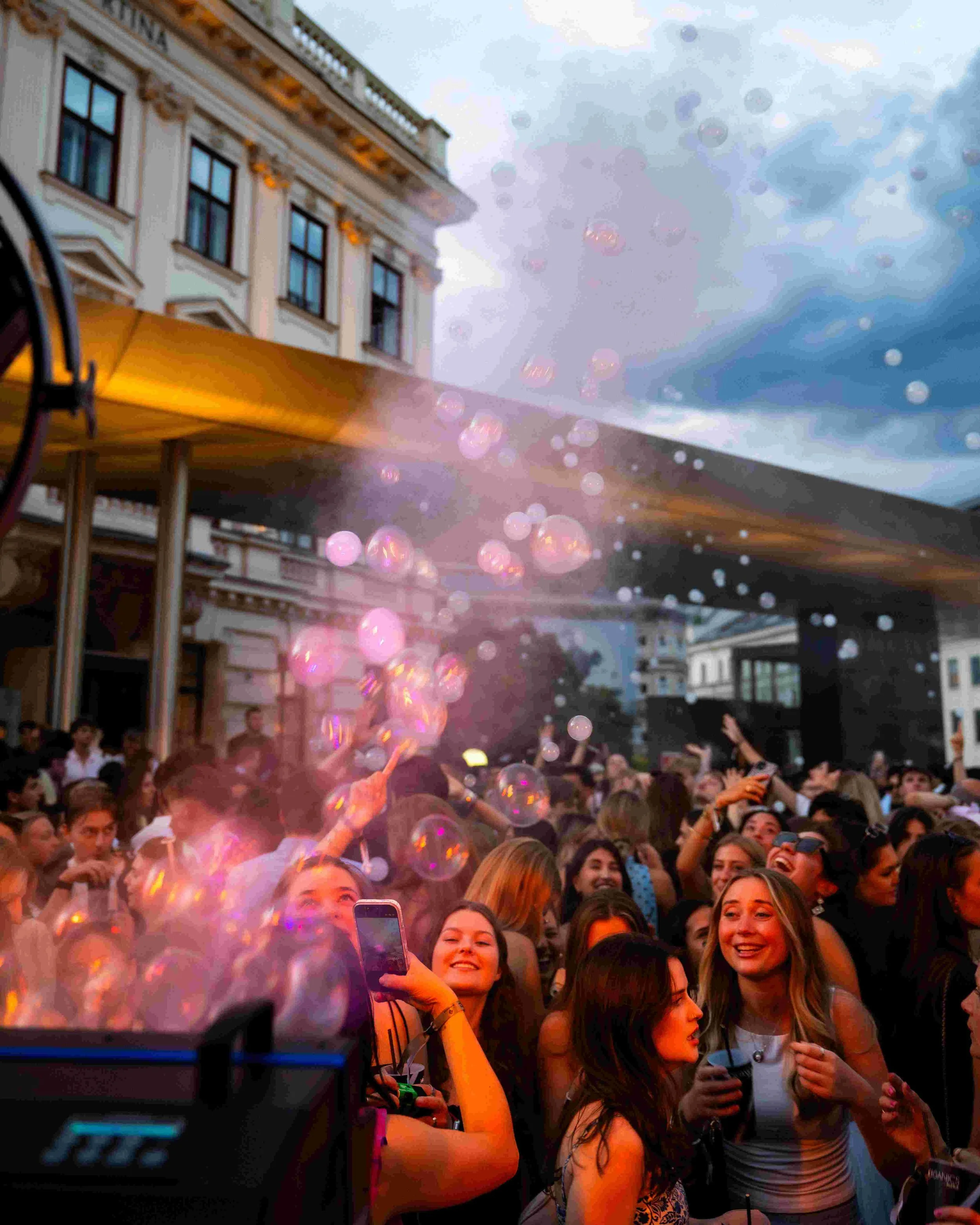 Crowd of excited young adults at an outdoor concert or festival, with balloons floating above and a building in the background.