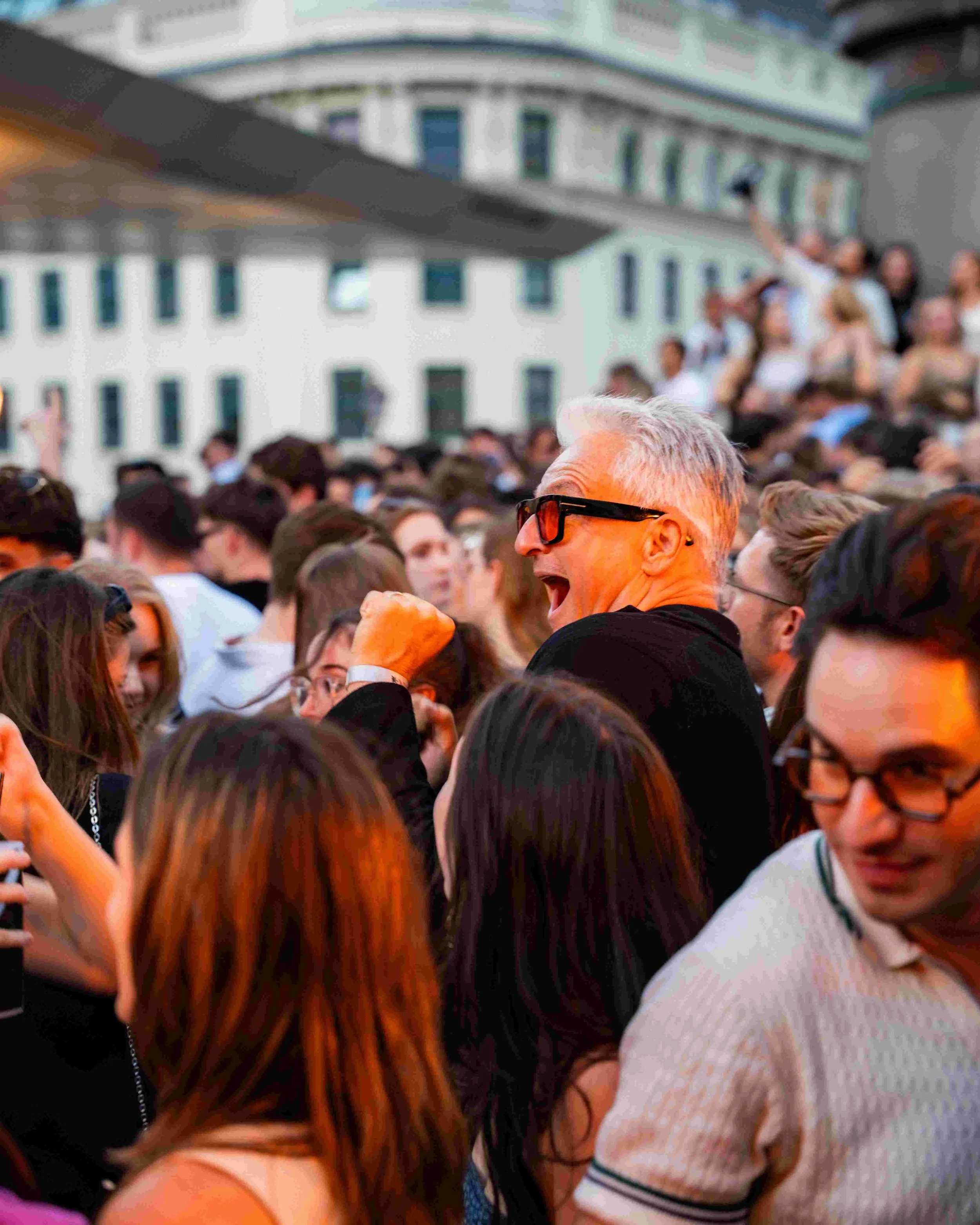 A crowd of people gathering outdoors with a large white building in the background, some individuals taking photos, and a man with gray hair and sunglasses in the foreground.