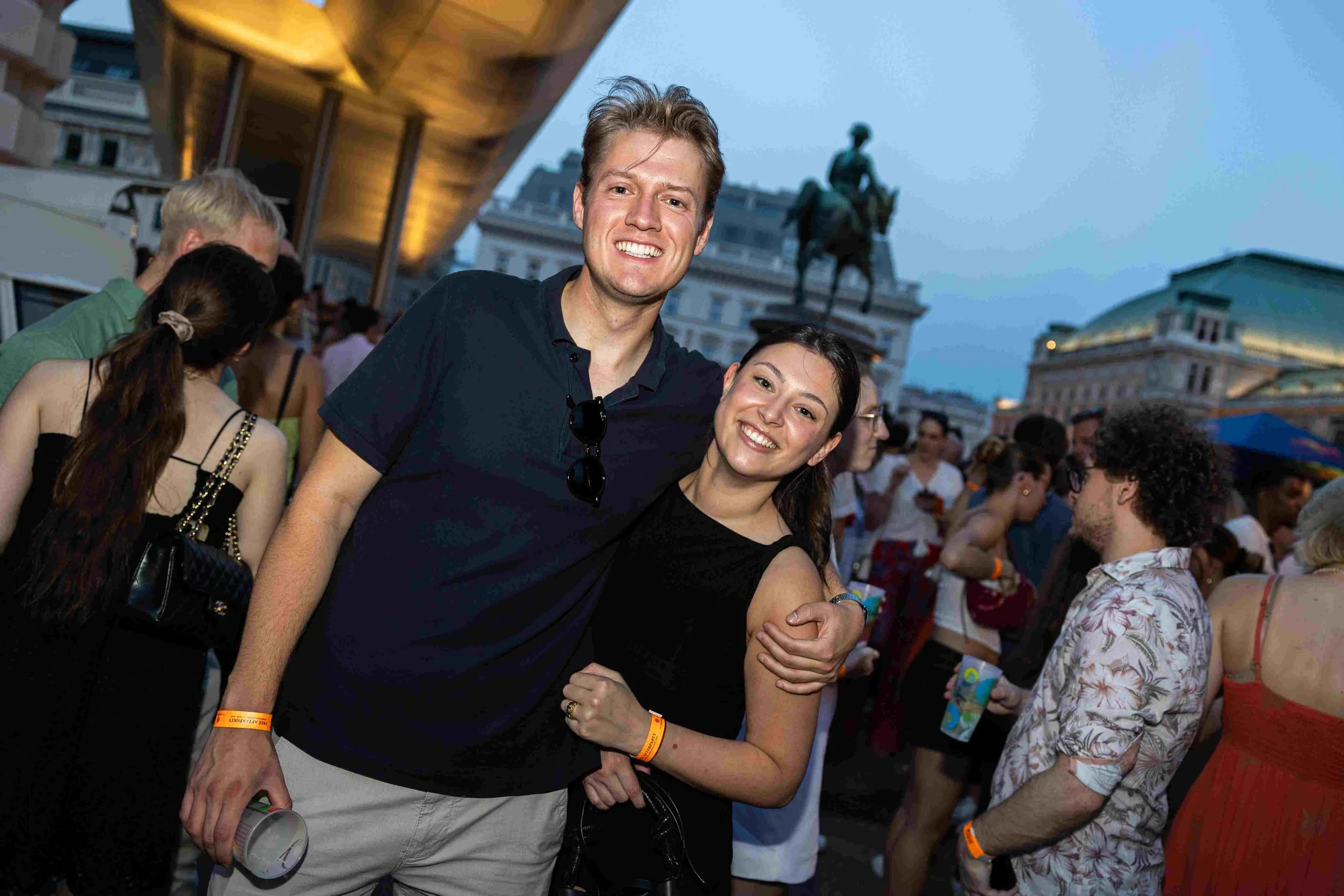 A smiling young man and woman embracing at an outdoor event in a city. There are many people around, some wearing festival wristbands, and background buildings with a statue of a mounted figure.