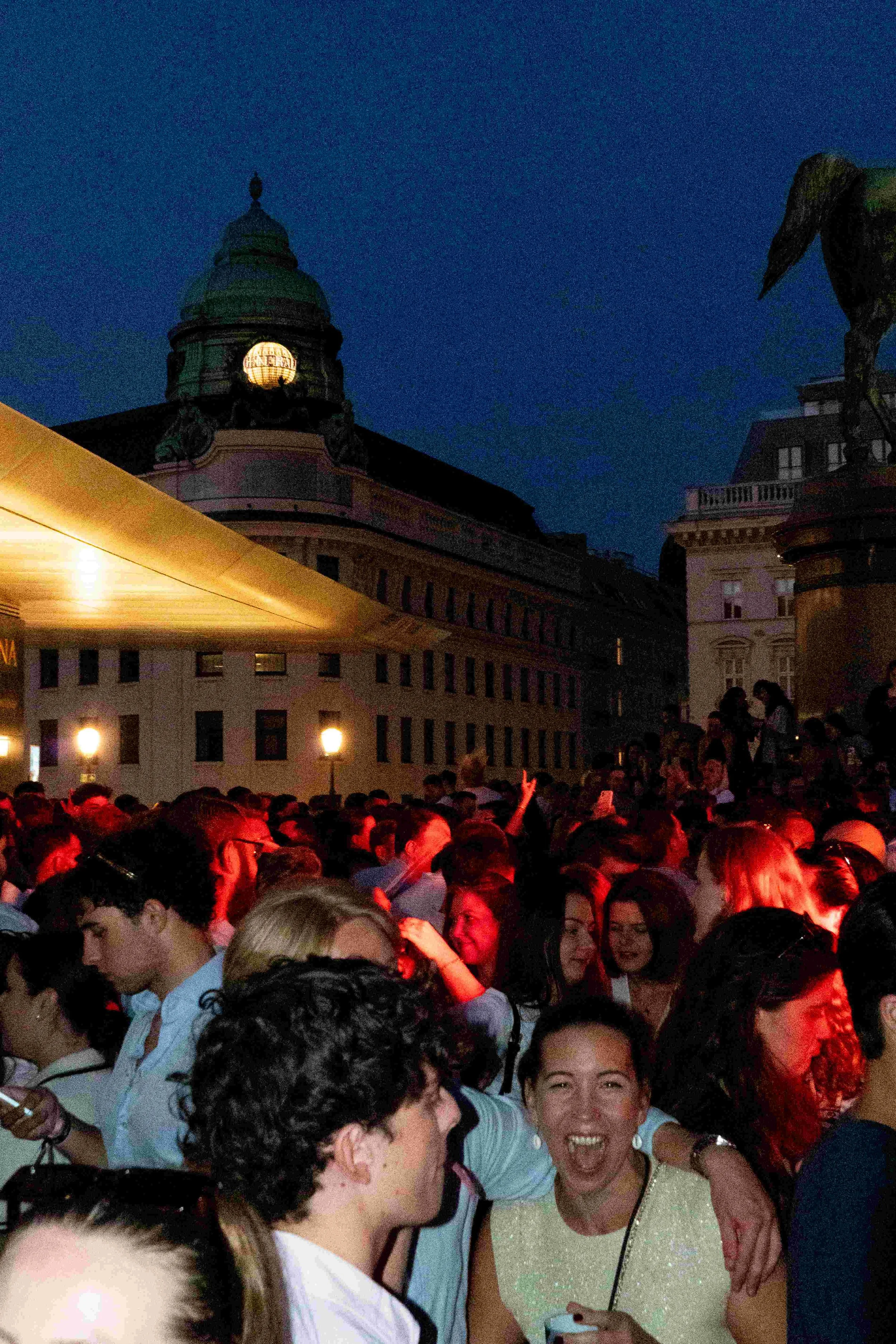 A large crowd of people gathered outdoors at night with a historic building and a statue of a horse in the background, illuminated by streetlights.