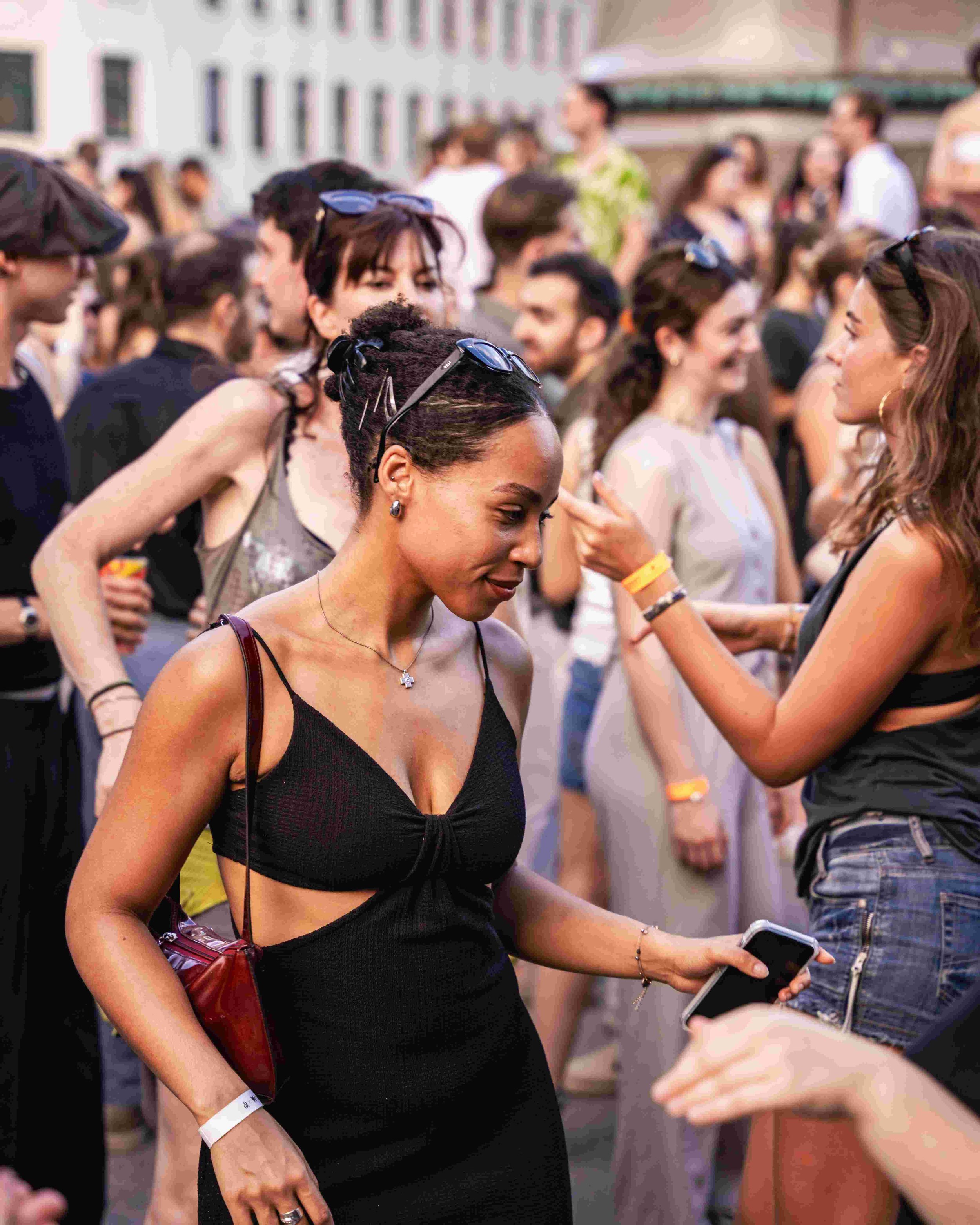 A diverse group of people gathered outdoors, socializing and engaging in conversations, with some holding smartphones. The scene appears to be at a social event or gathering.