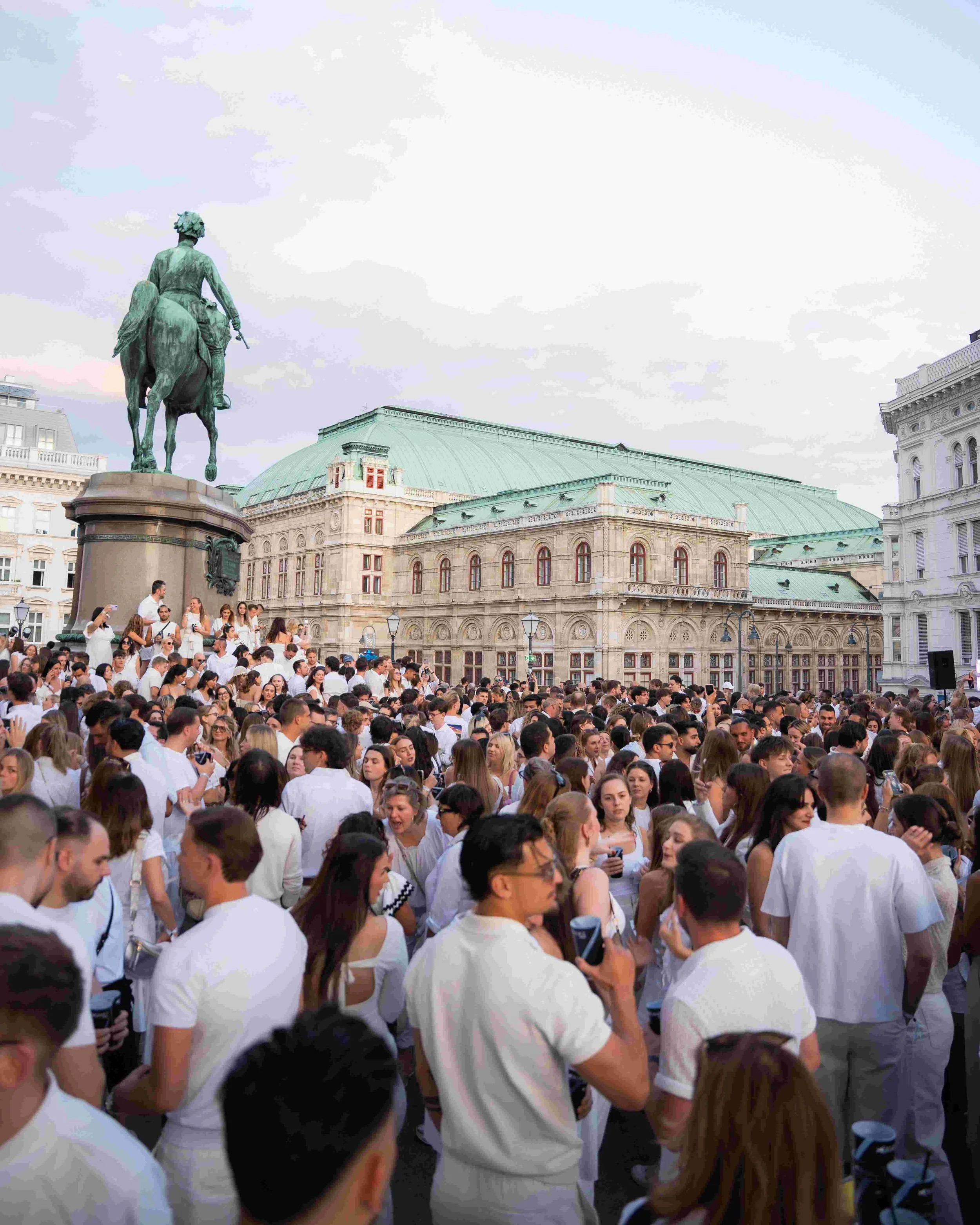 Crowd of people wearing white shirts, some taking photos, gathered in a city square with historic buildings and a prominent equestrian statue of a man on horseback in the background.
