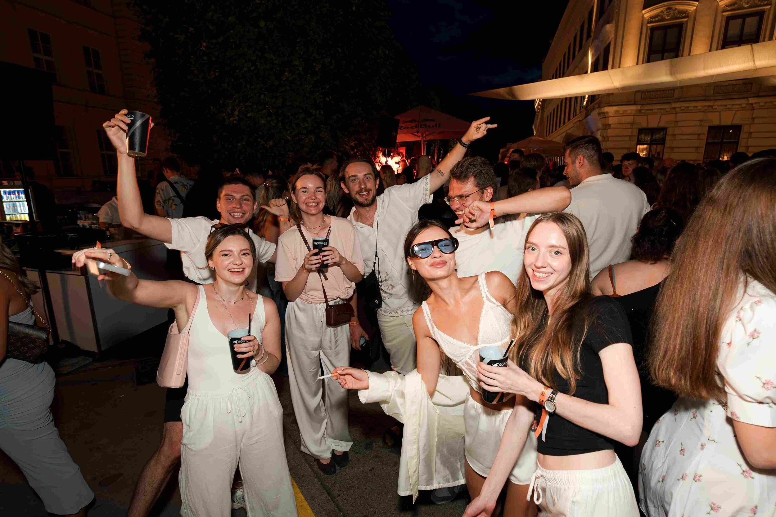 Group of young adults enjoying a night out at an outdoor event, smiling and holding drinks, with a DJ and stage in the background.