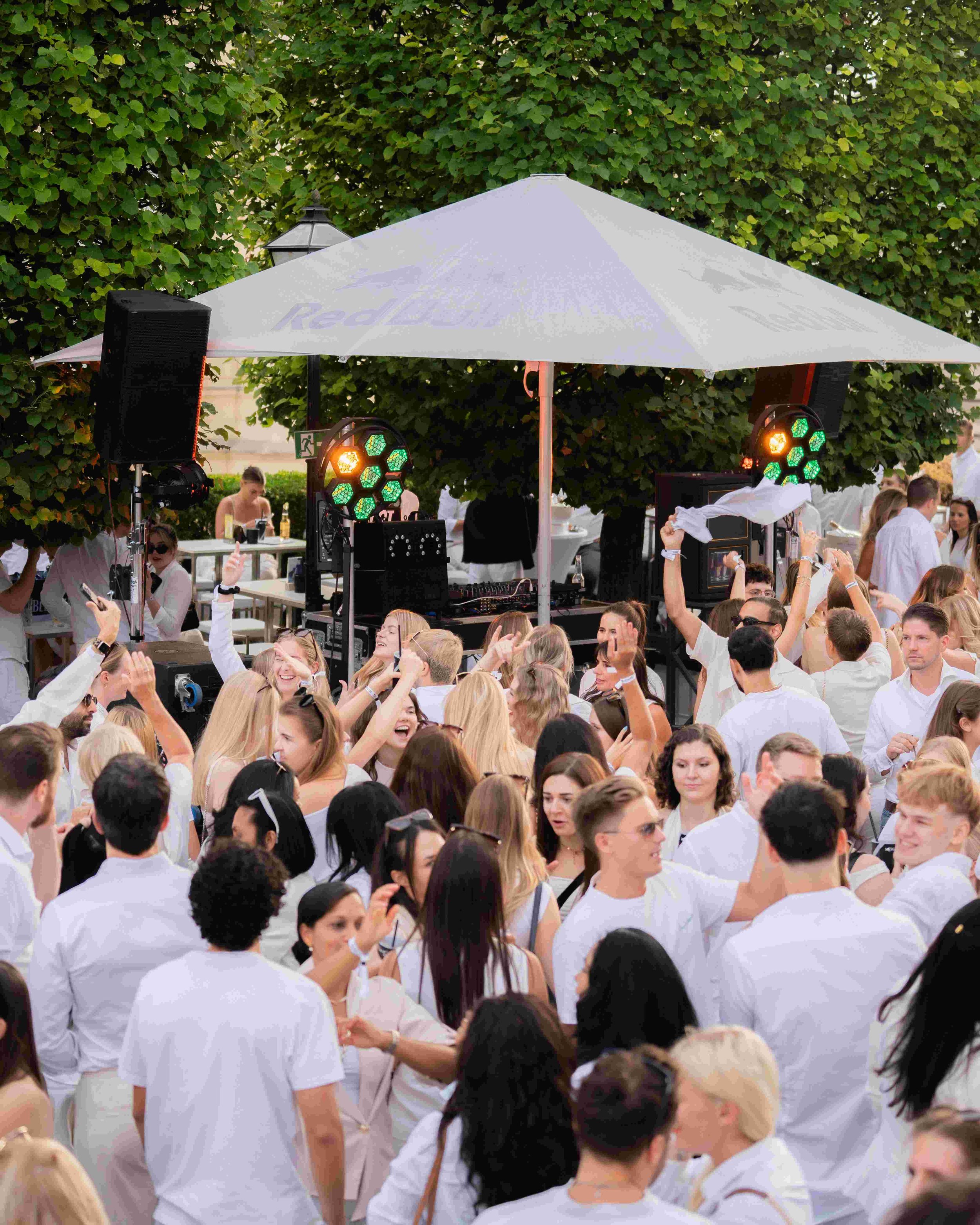 Crowd of people gathered outdoors under a large white umbrella with green trees in the background, enjoying a social event.