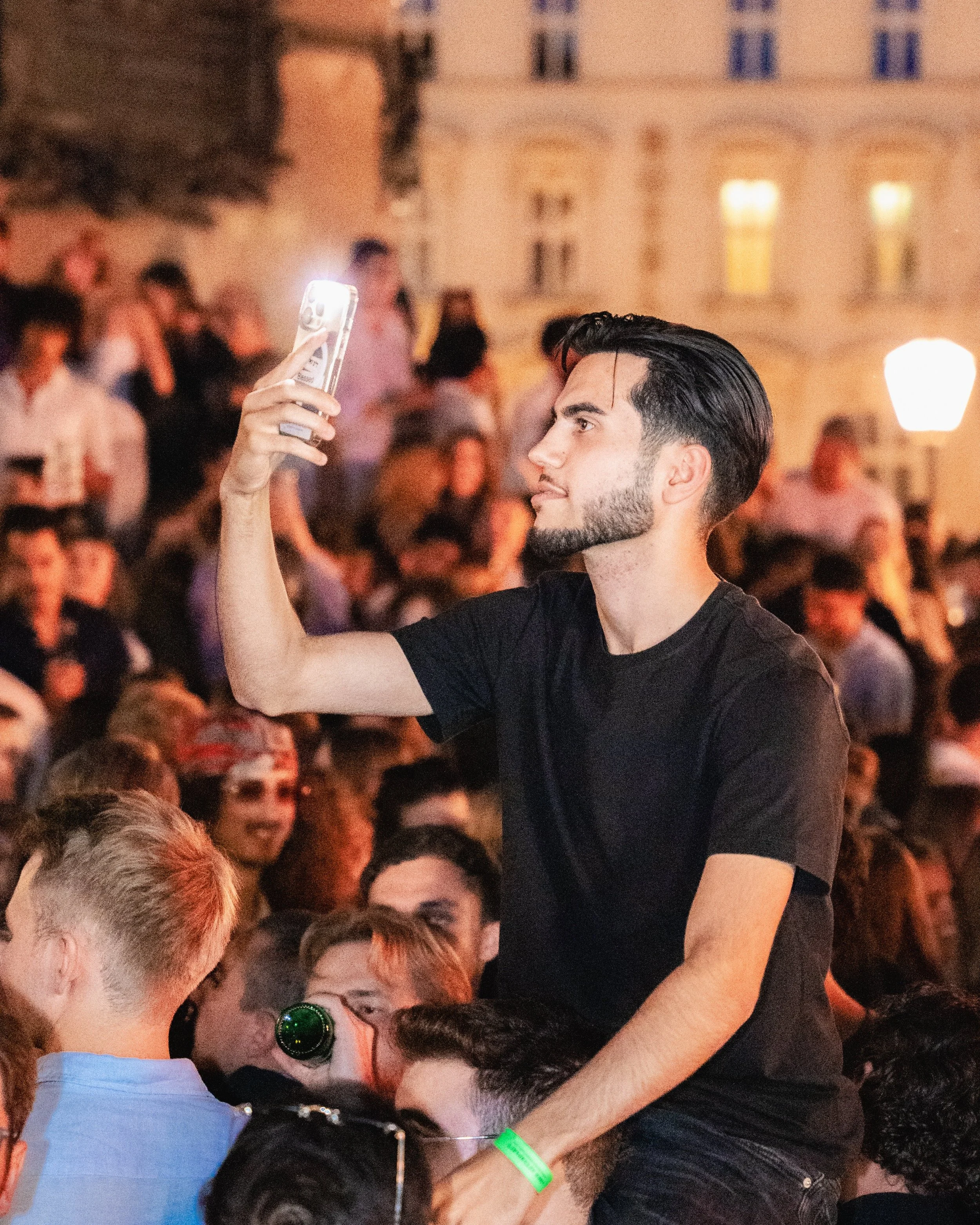 A young man with dark hair and beard taking a selfie with a smartphone at a nighttime event surrounded by many people.