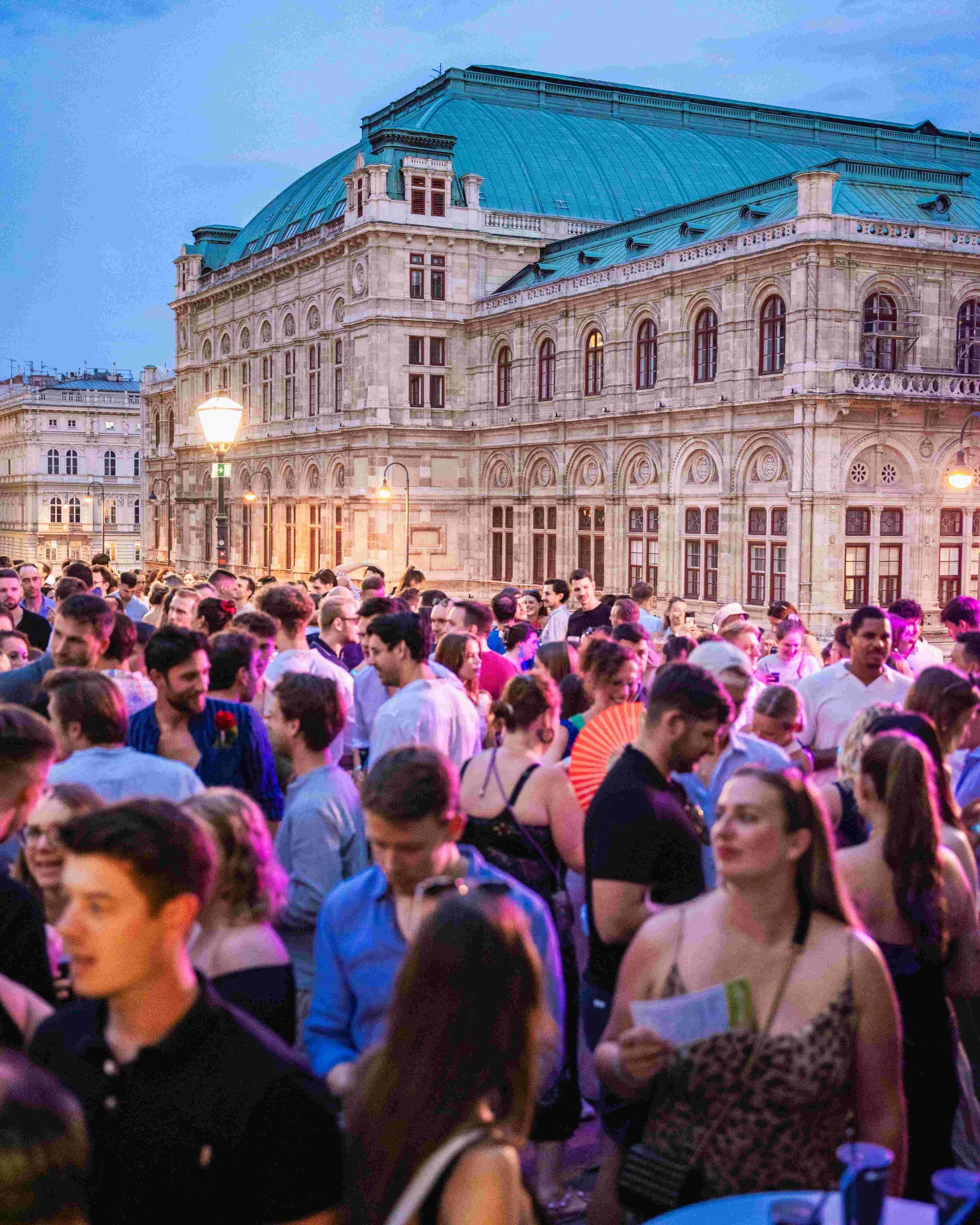Crowd of people gathered outdoors in front of historic building with ornate architecture and green domed roof during evening.