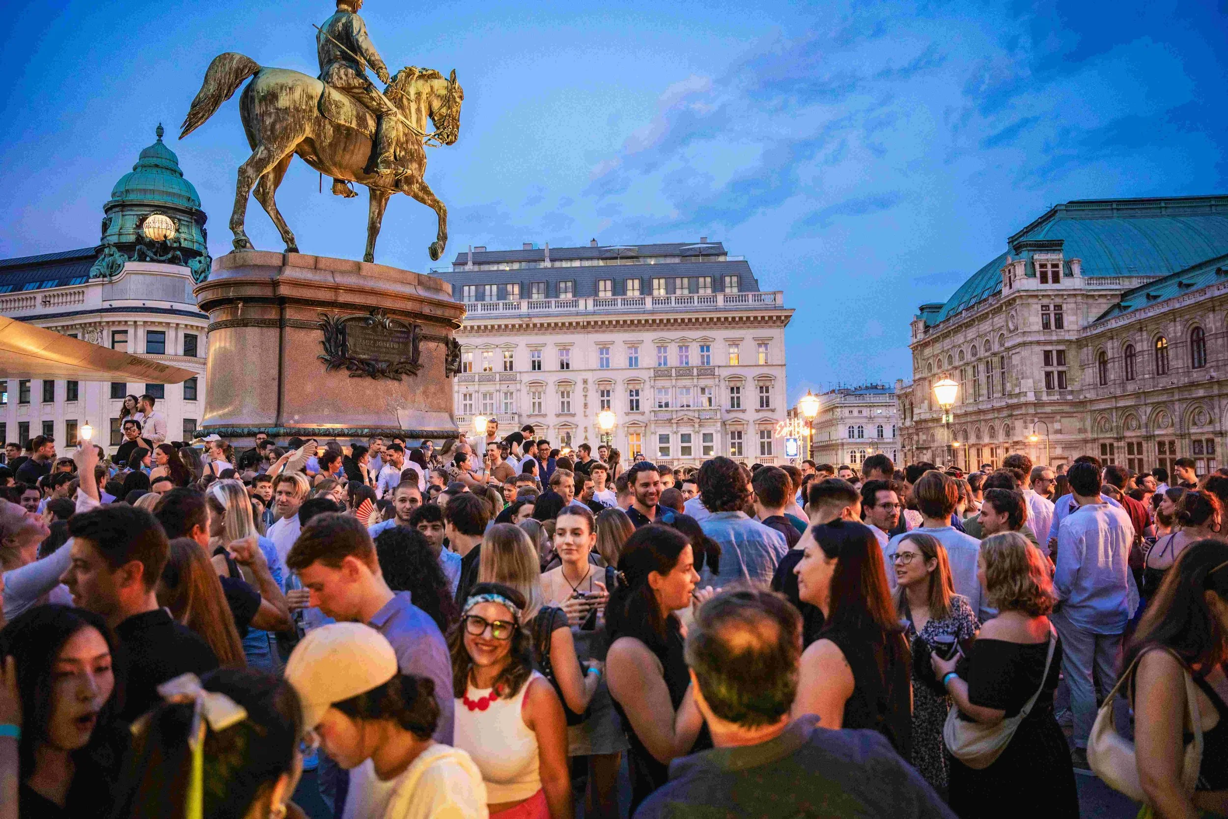Large crowd of people gathered outdoors in front of a statue of a man on a horse, in an urban setting during the evening. The scene includes historic buildings with European architectural style and street lamps lit, with a partly cloudy sky in the ba
