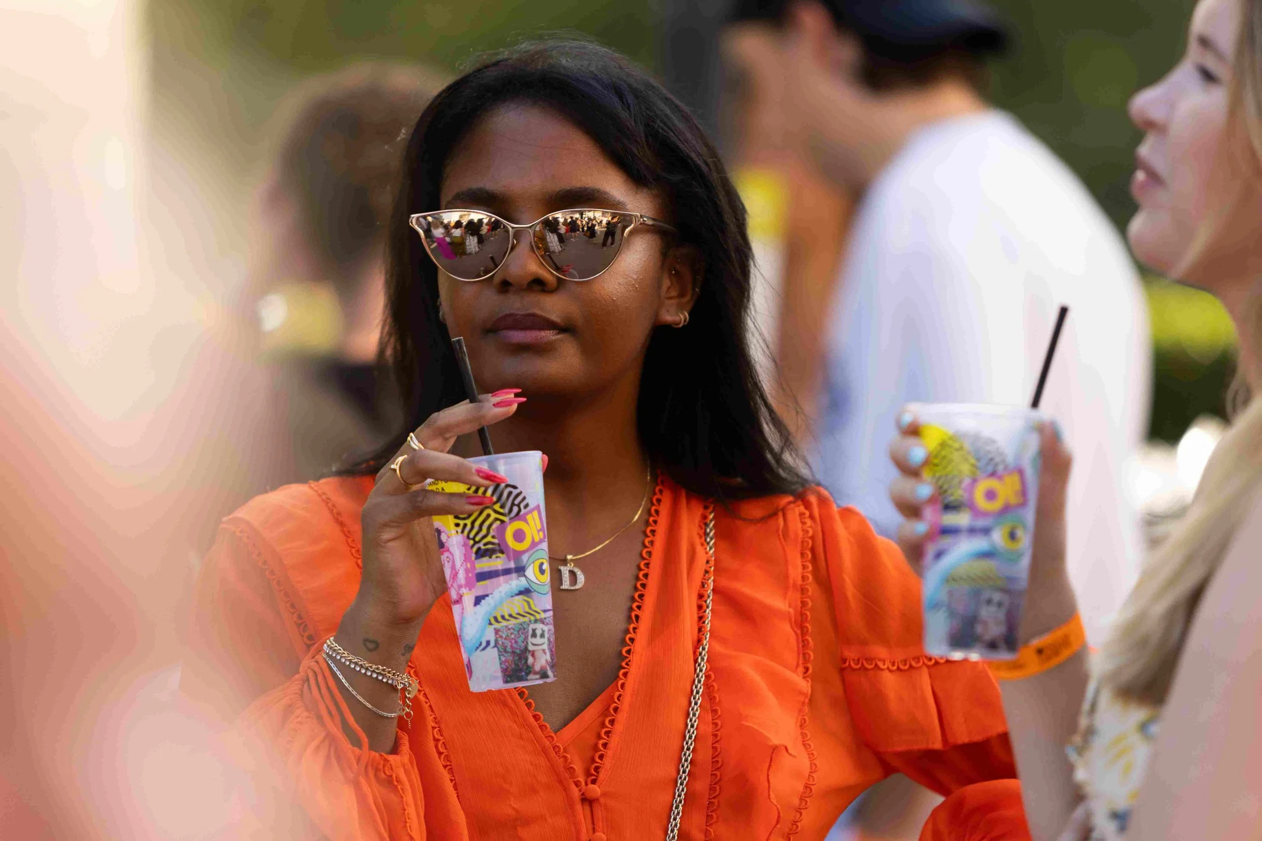 Woman with black hair wearing sunglasses and orange clothing holding a colorful ice drink at an outdoor event.