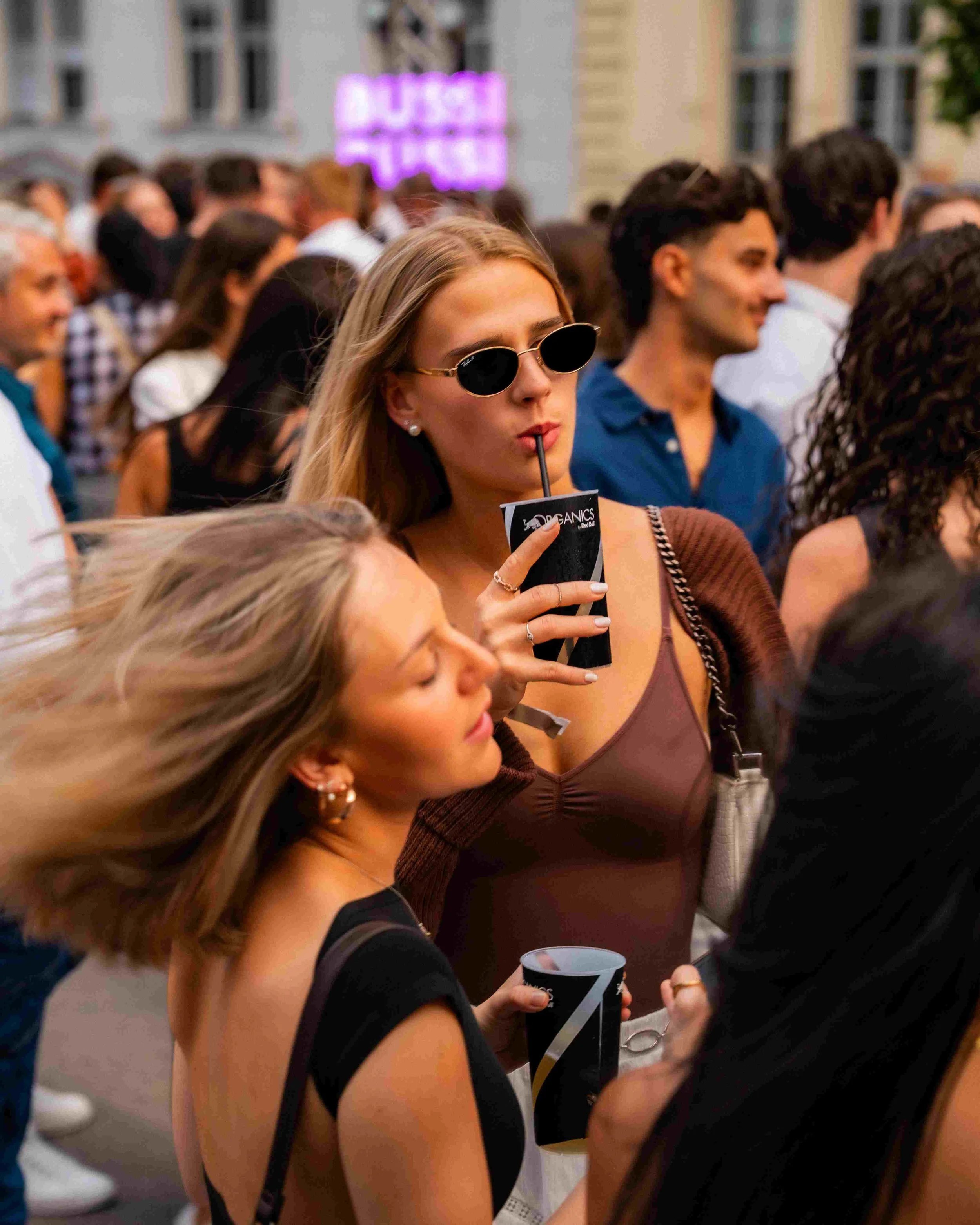 A group of people gathered at an outdoor event, with a young woman in the foreground sipping a drink through a straw while wearing sunglasses and holding a cup with a logo. The background shows other attendees and a building with pink neon sign.