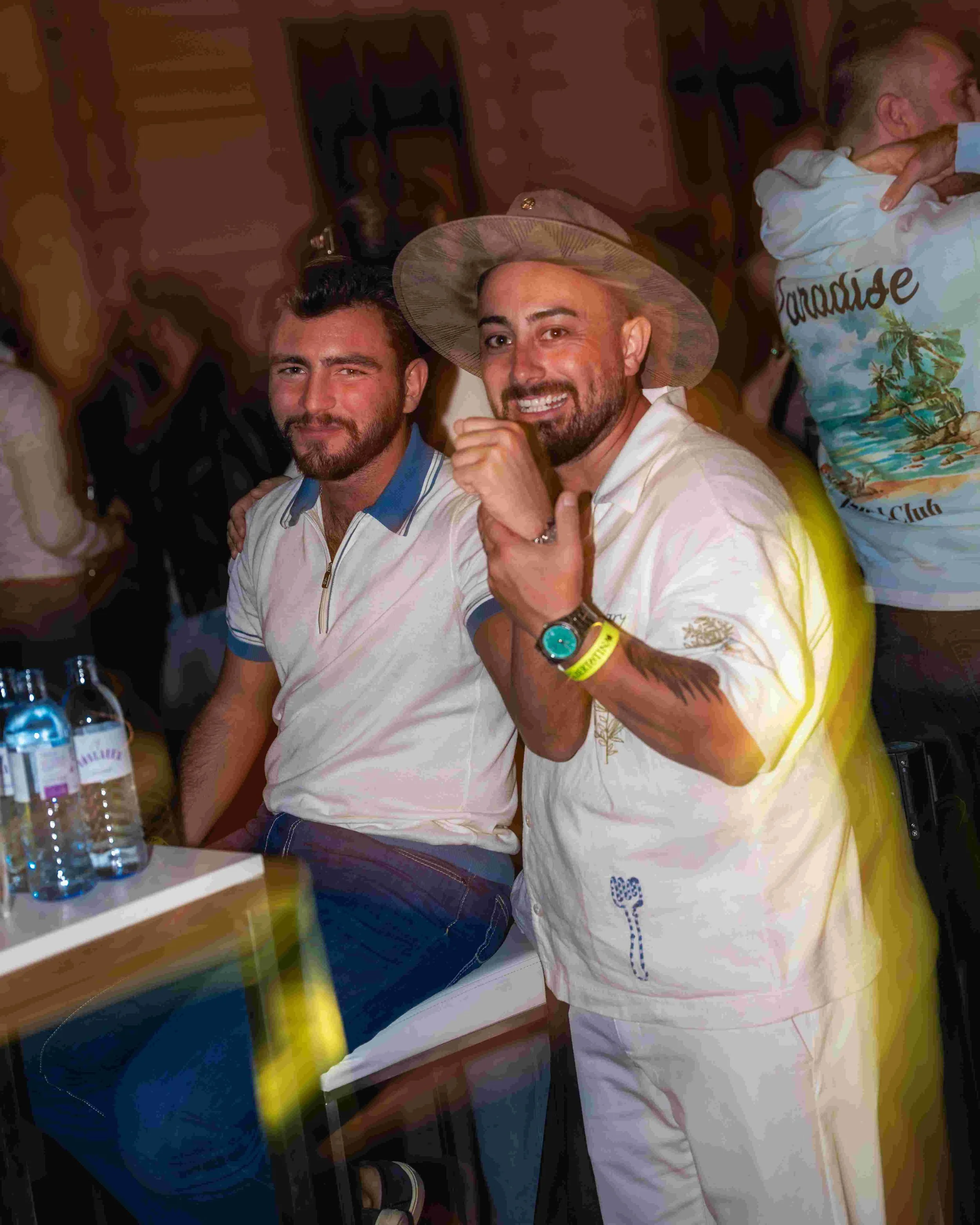 Two men sitting at a bar or restaurant, smiling at the camera. The man on the left has dark hair and a beard, wearing a short-sleeved shirt with blue collar and striped trim. The man on the right has light hair, a beard, and is wearing a wide-brimmed