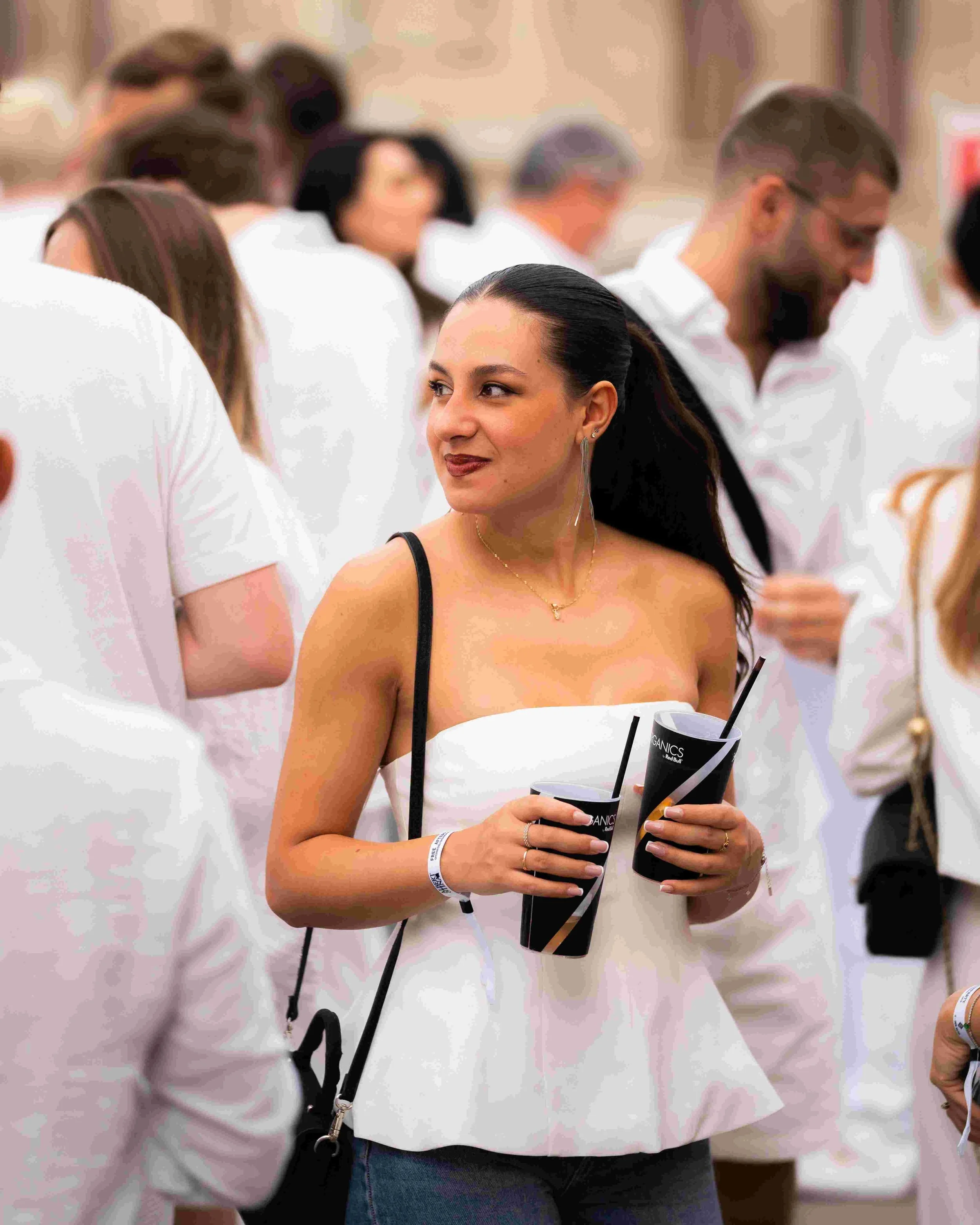 A woman with dark hair in a ponytail, wearing a strapless white top, holding a black cup with a straw, at a crowded event with people standing around.