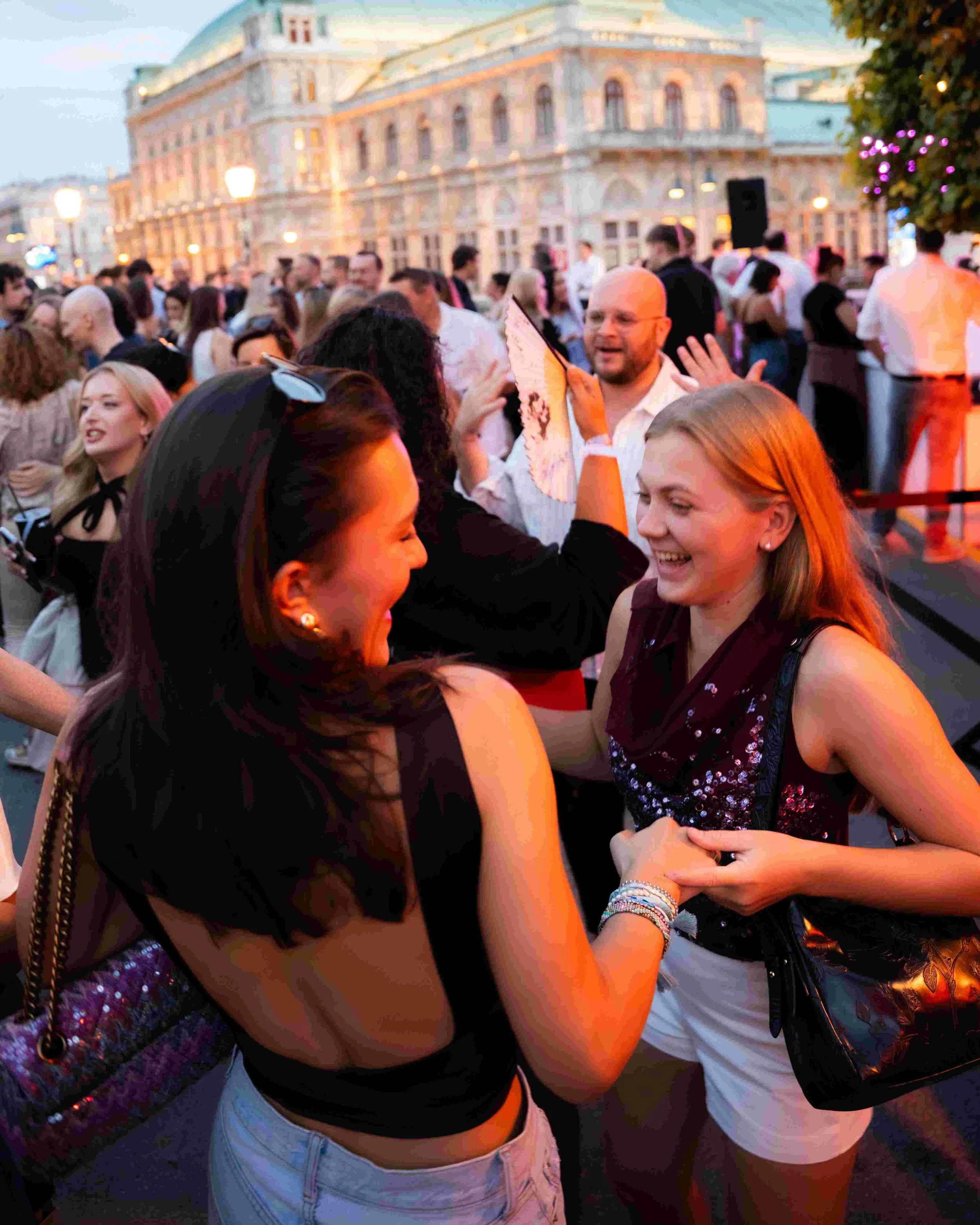 Group of young people dancing and socializing at an outdoor event in the evening with historic buildings and streetlights in the background.