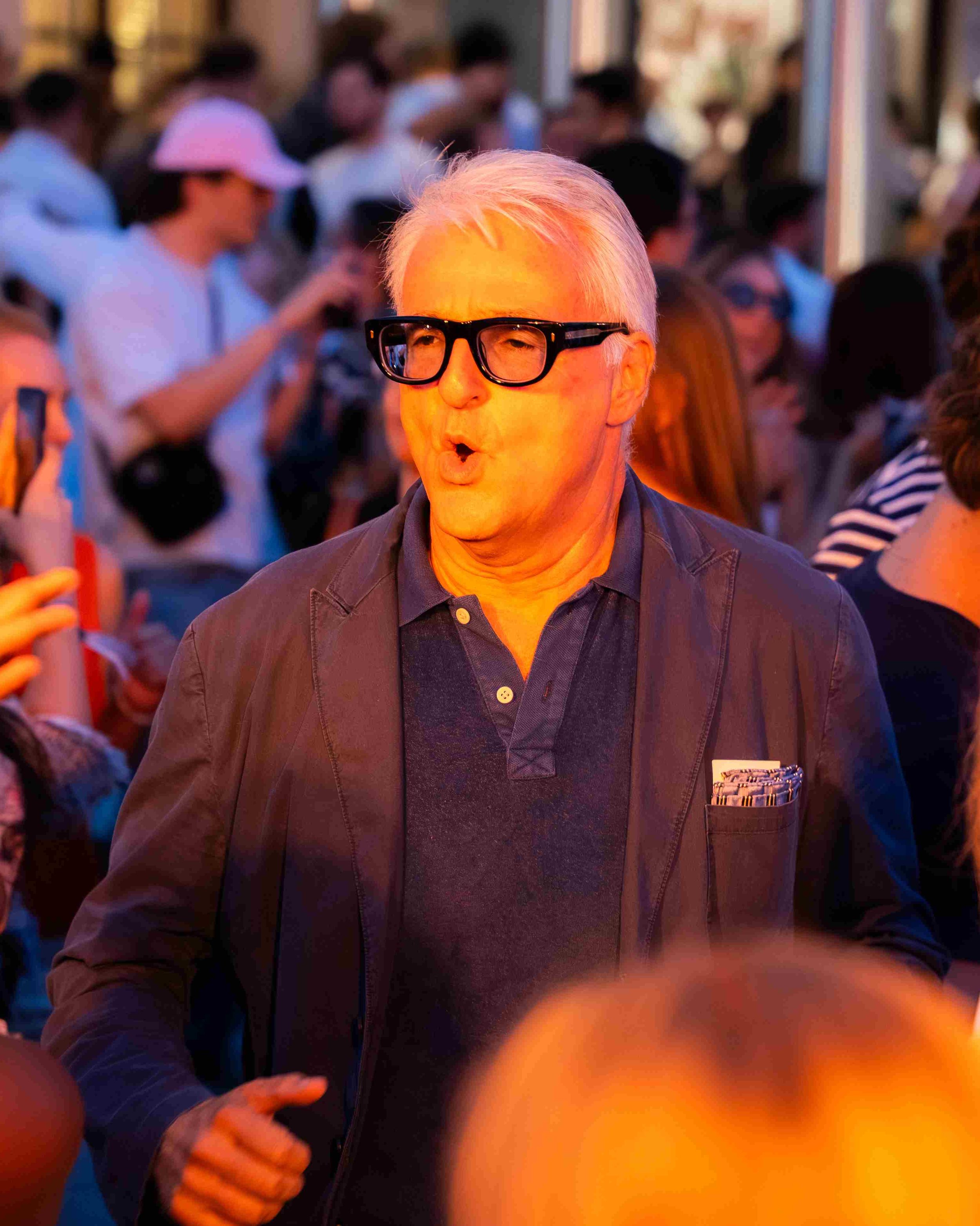 A man with white hair and black glasses is in the foreground at a crowded outdoor event, making a surprised or expressive face. Multiple people are in the background, some wearing sunglasses and hats.
