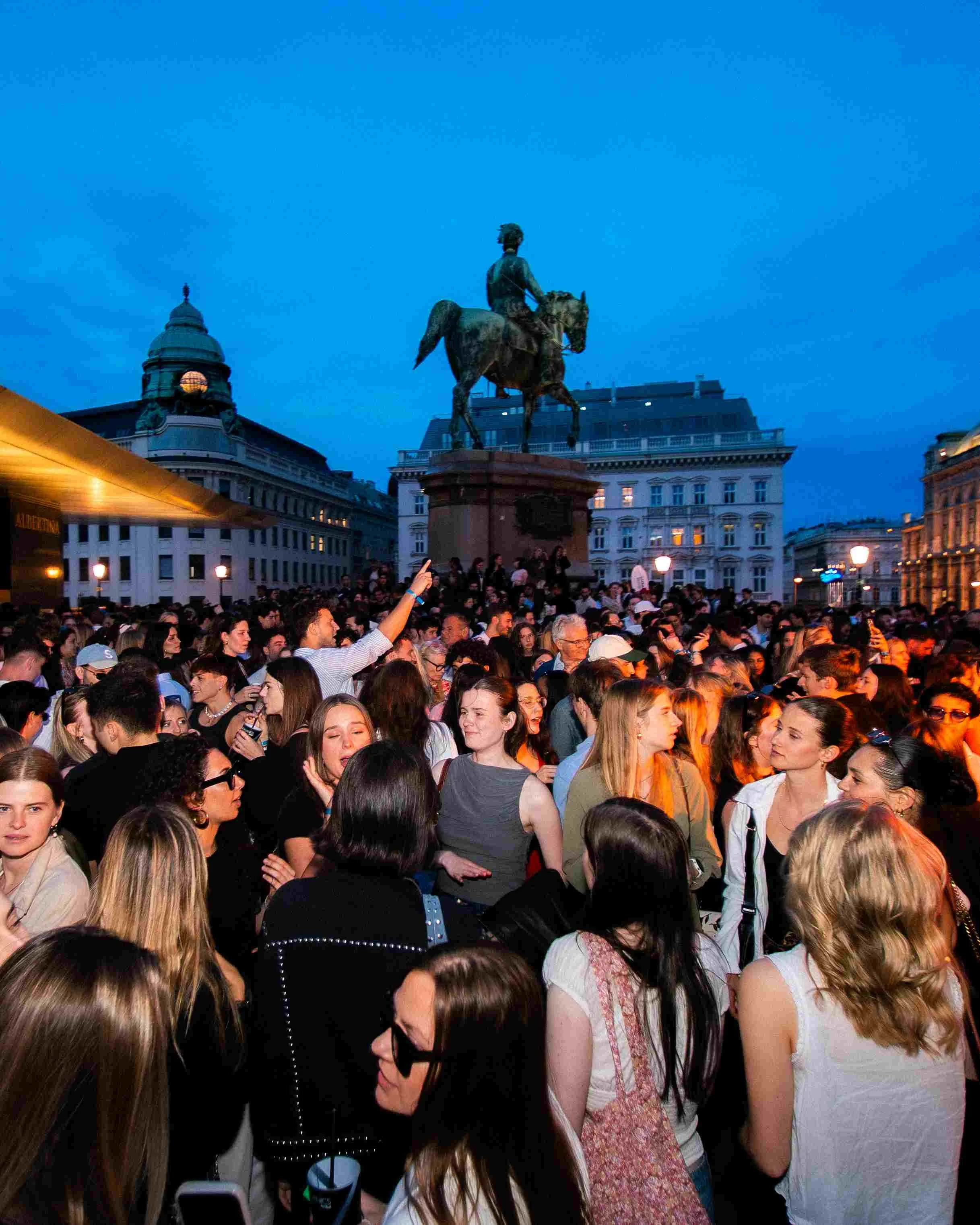 A large crowd of people gathered outdoors in an urban area during evening, with some holding up devices and taking photos. In the background, there is an equestrian statue of a man on a horse, situated on a city square with historic buildings around.