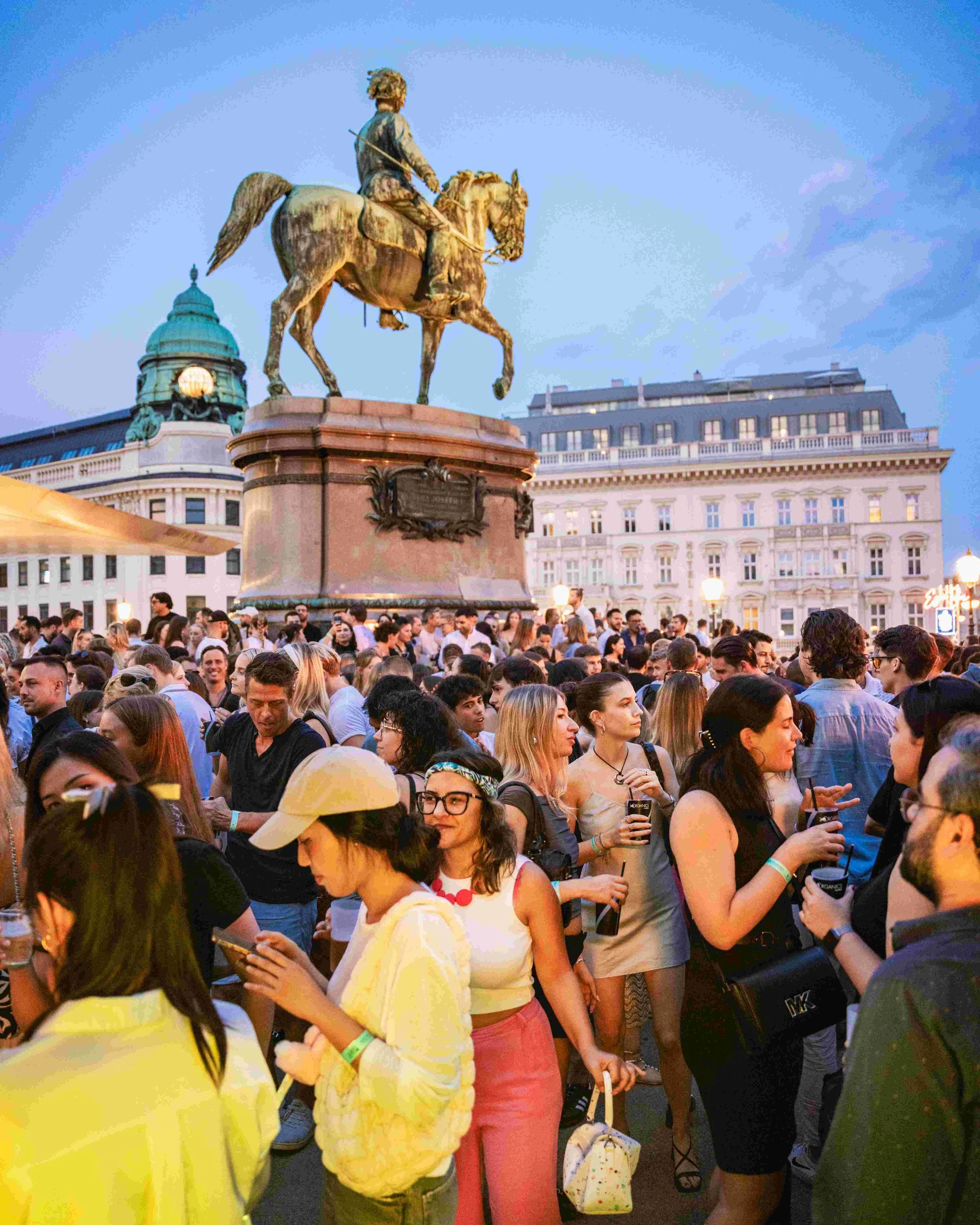 Crowd of people gathered outdoors during evening, with an equestrian statue in the background and historic buildings with illuminated windows.
