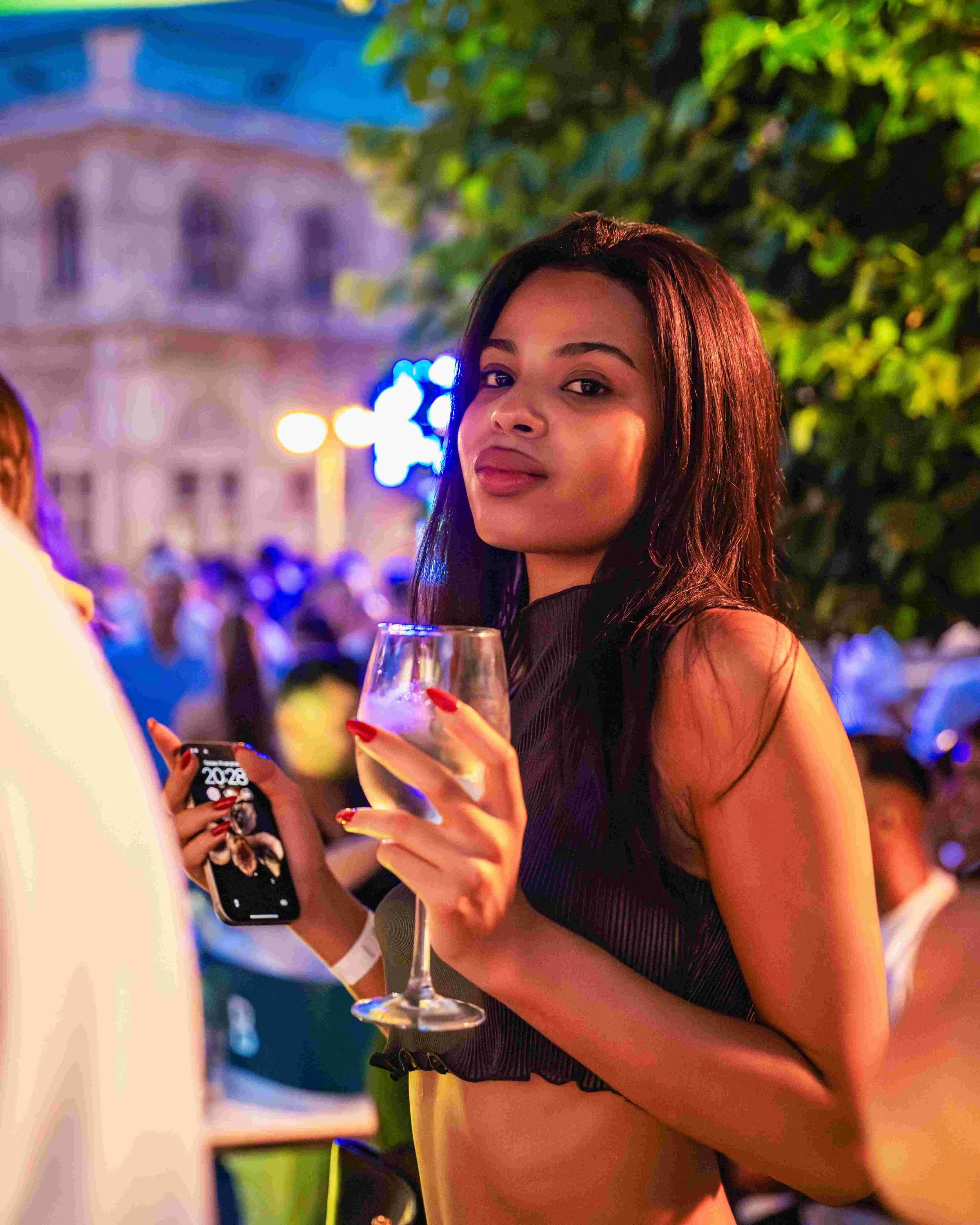 A young woman with long brown hair and light brown skin holds a glass of white wine in her right hand at an outdoor event during the evening, with vibrant blue and yellow lighting and an out-of-focus building in the background.