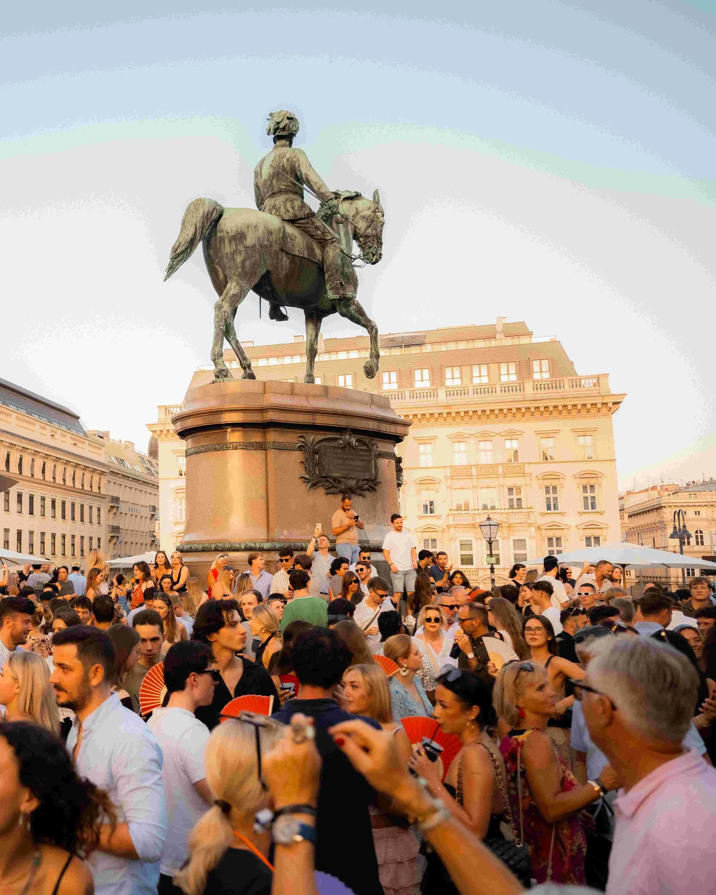 A crowd of people gathered in a city square with a large equestrian statue and historic buildings in the background during daytime.