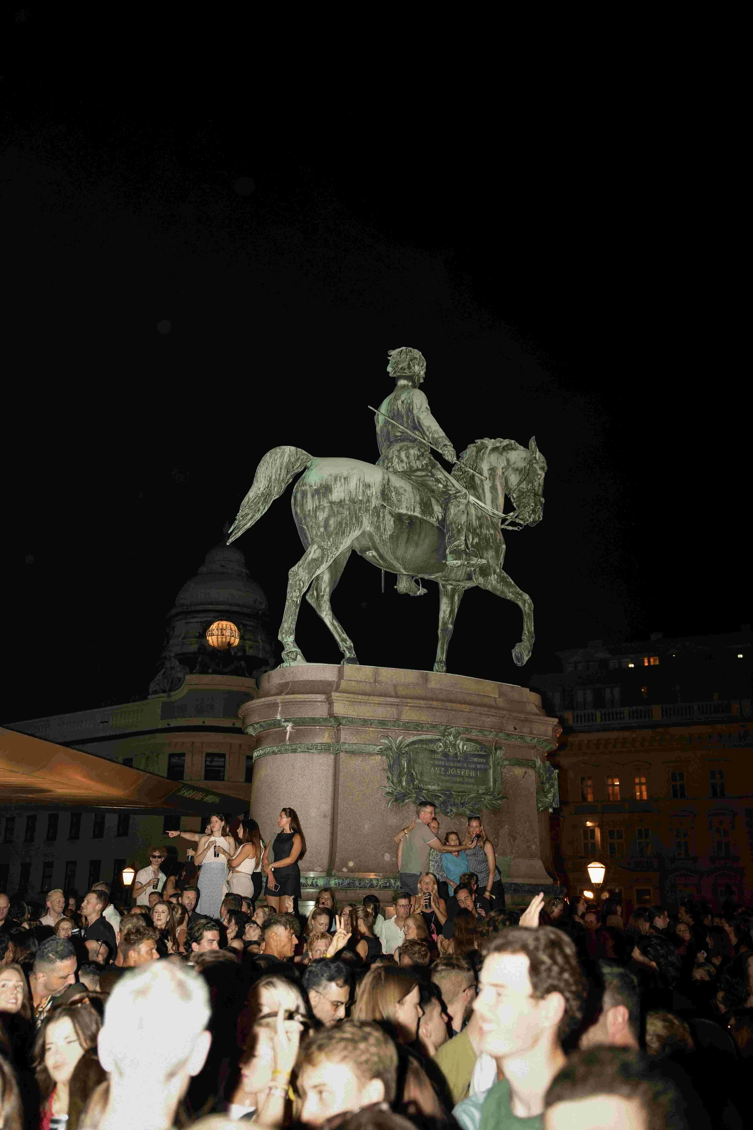Nighttime scene with an equestrian statue of Franz Joseph, surrounded by a large crowd of people, some taking photos, in an urban square.