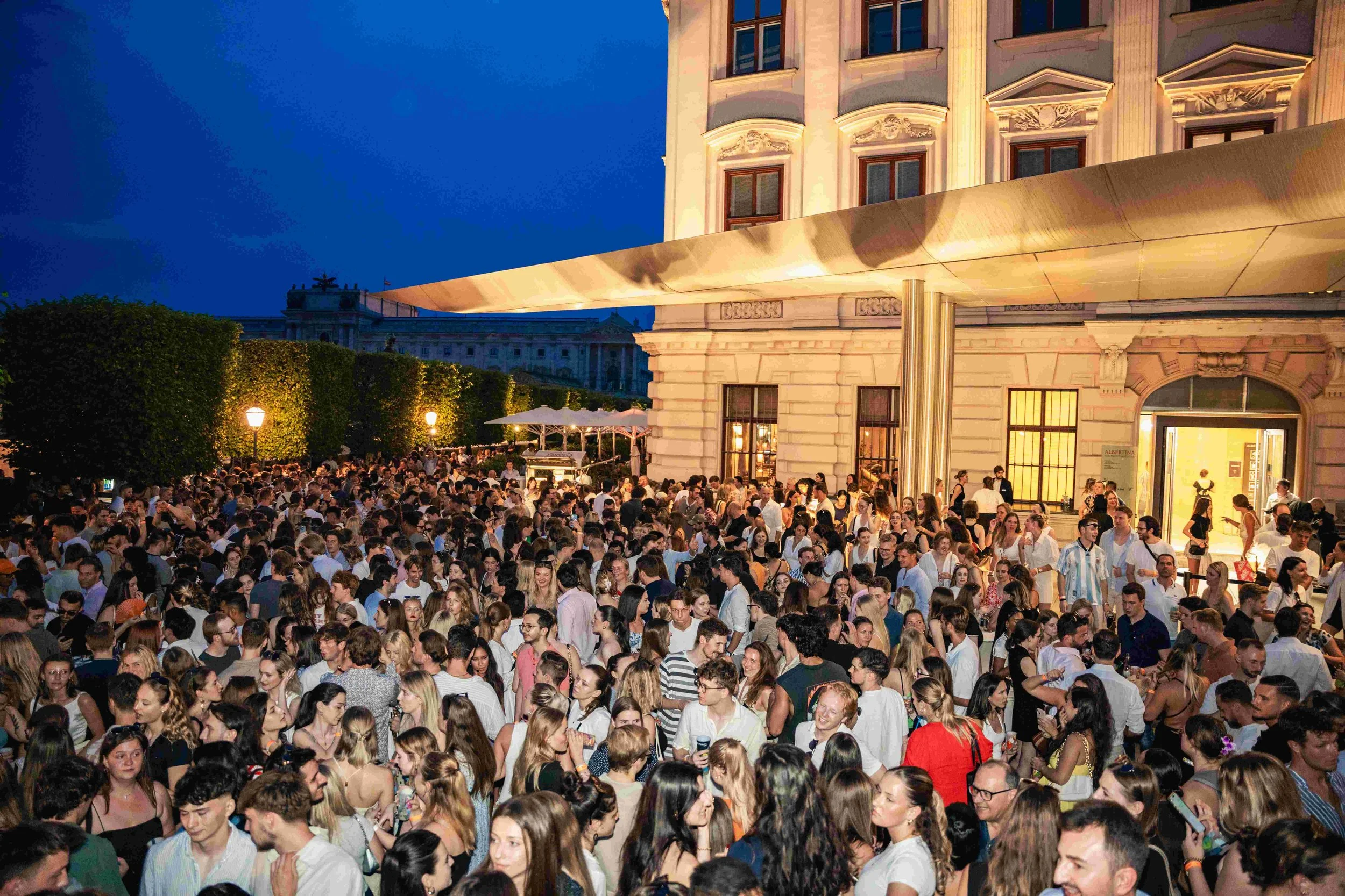 A large gathering of people outside a historic building at dusk, with trees and street lamps visible, and a modern architectural awning over the entrance.
