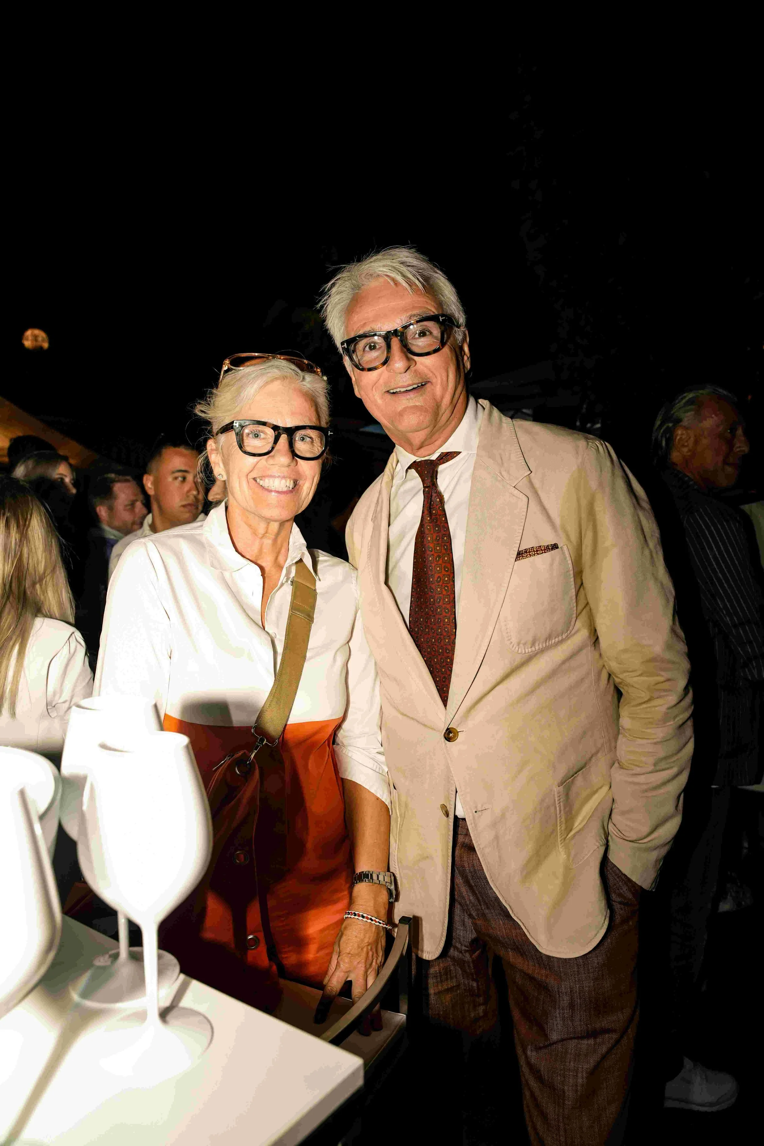 A smiling older woman and man wearing glasses and dressed in business casual attire standing close together at a social event, with people in the background.