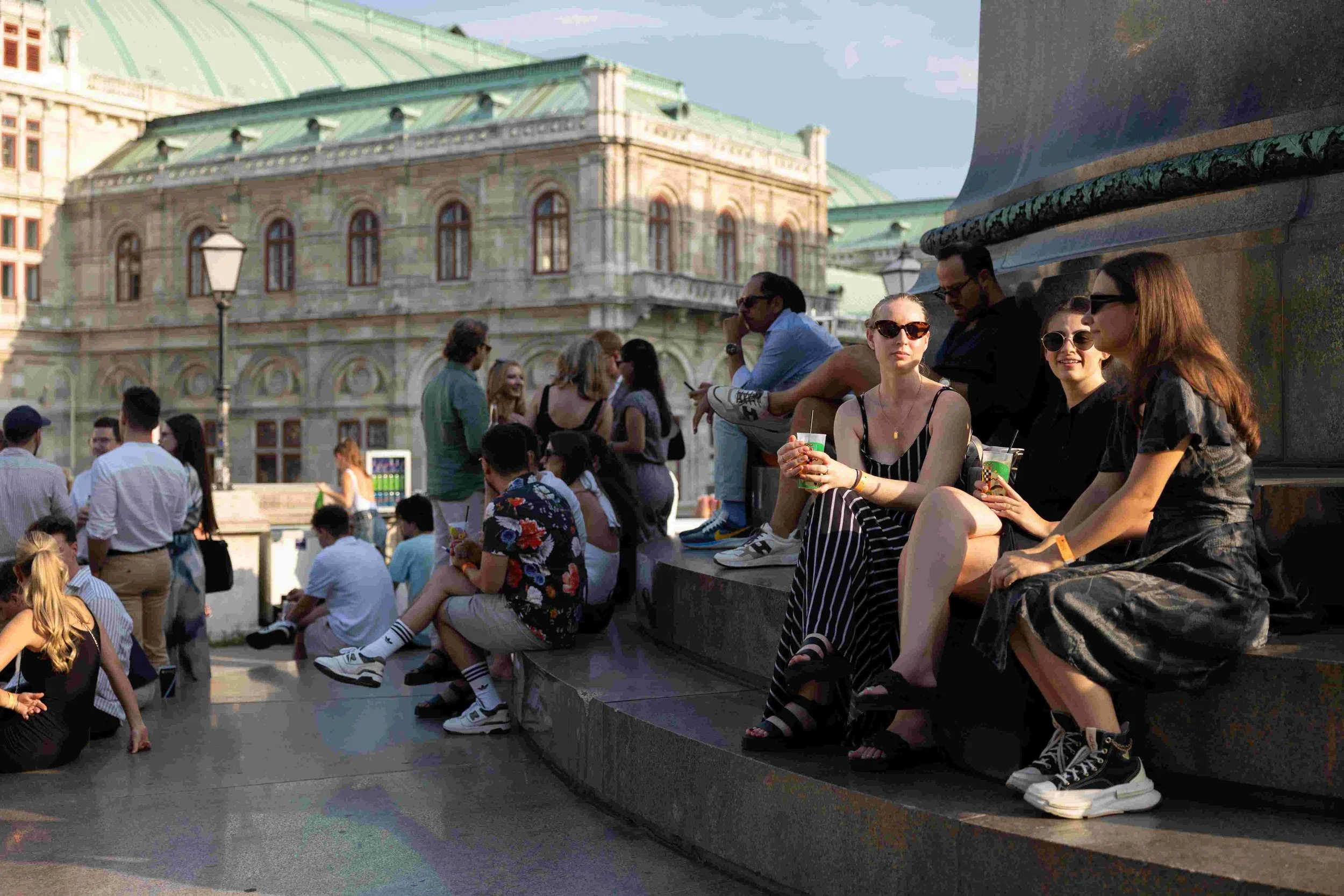 People sitting on a ledge in a city square, engaging in conversation and enjoying the weather, with historic buildings in the background.
