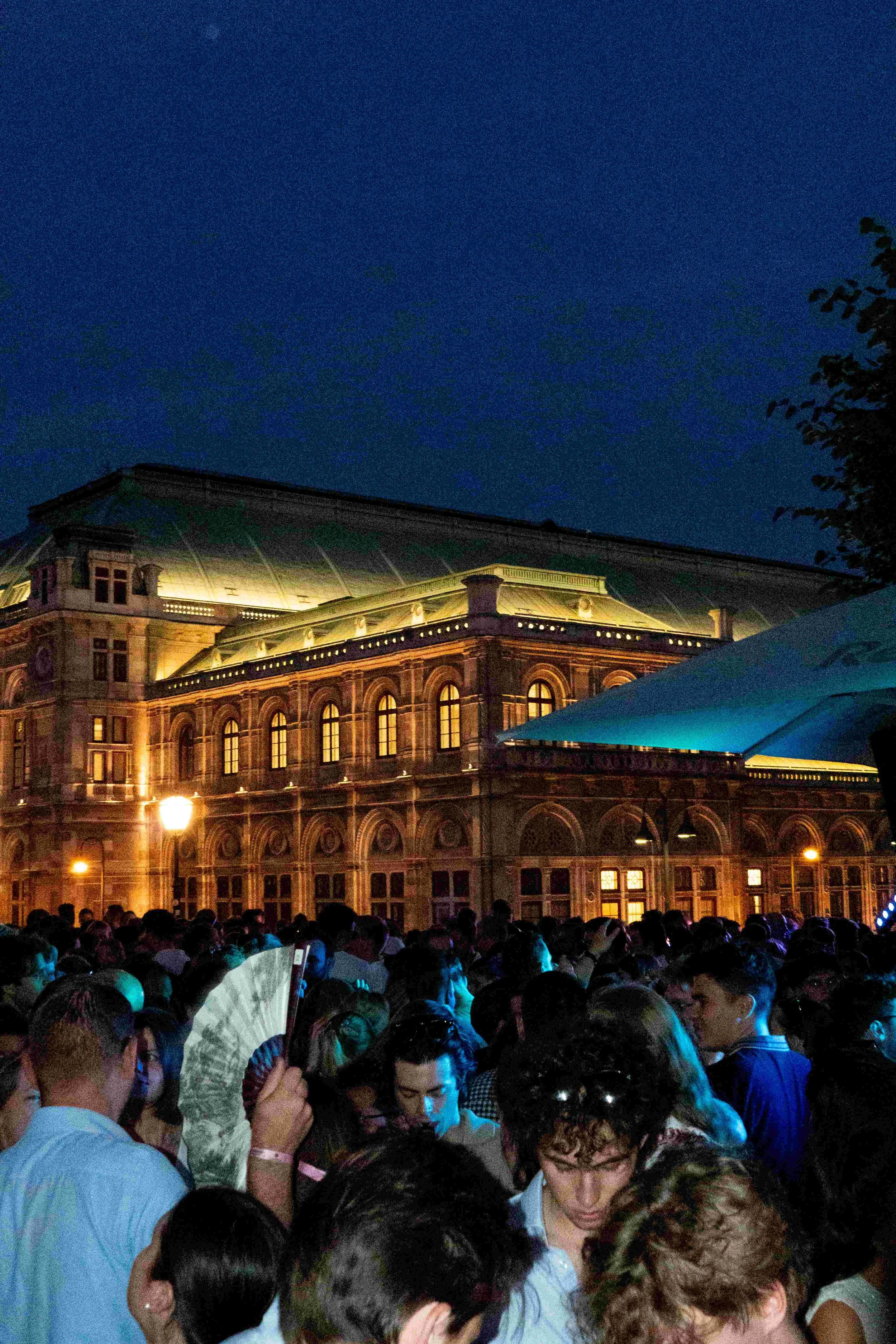 A large crowd of people gathered outside a historic building at night, illuminated by warm lights, with a clear dark blue sky overhead.