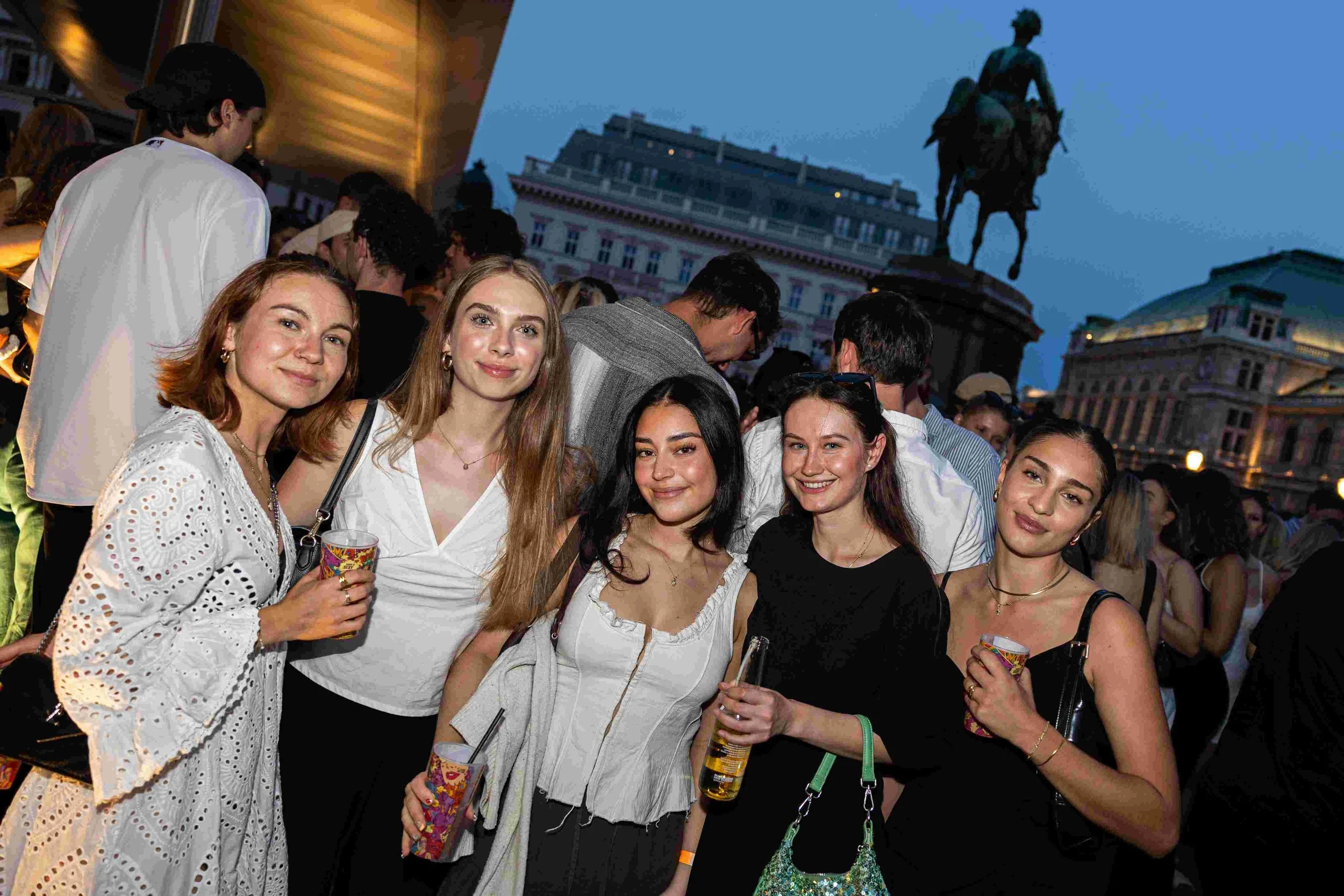 A group of young women smiling and holding drinks at an outdoor evening event, with an equestrian statue and city buildings in the background.