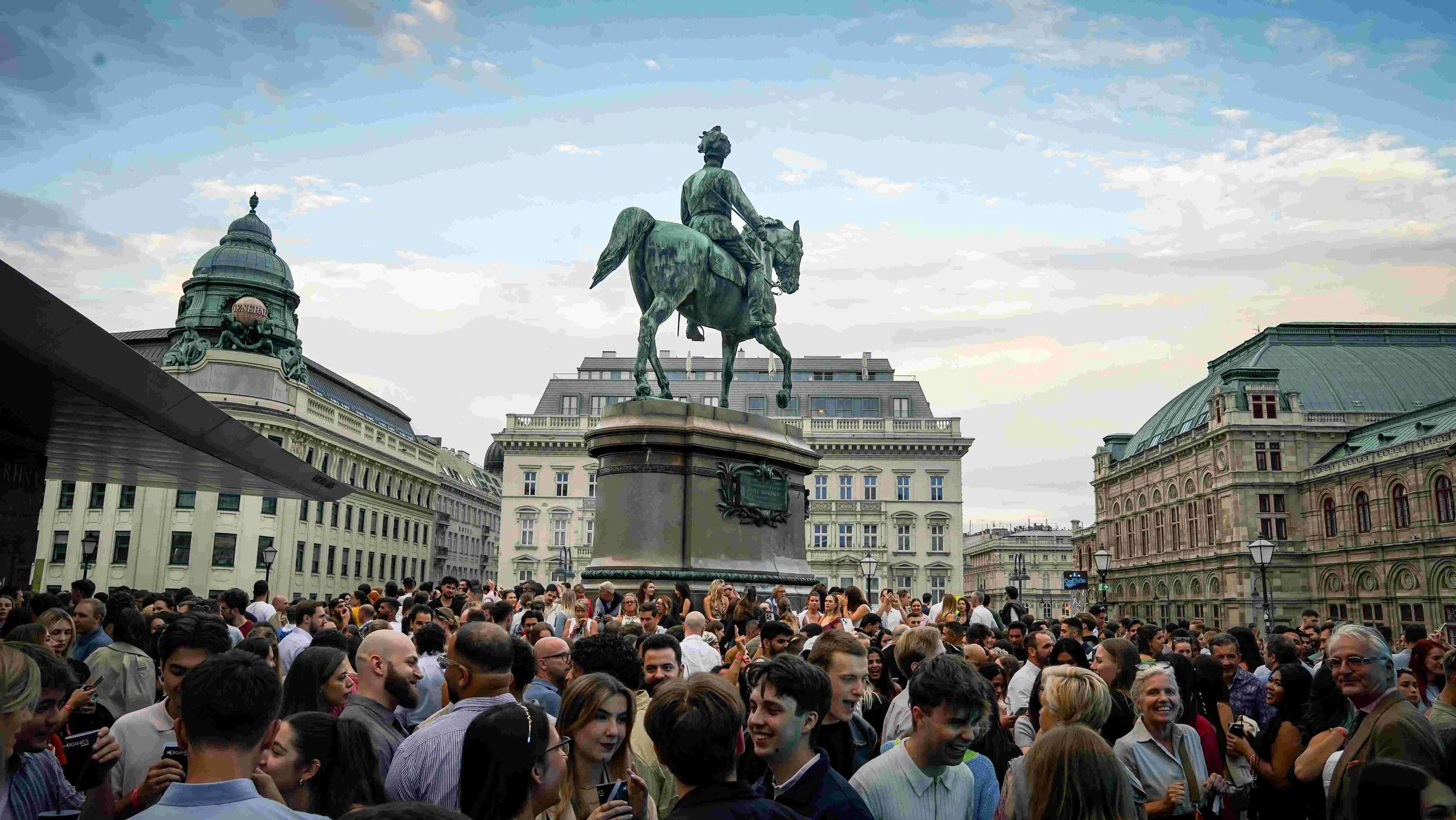 Crowd of people gathered in an urban square with historic buildings, a prominent statue of a man on a horse in the center, and a partly cloudy sky overhead.