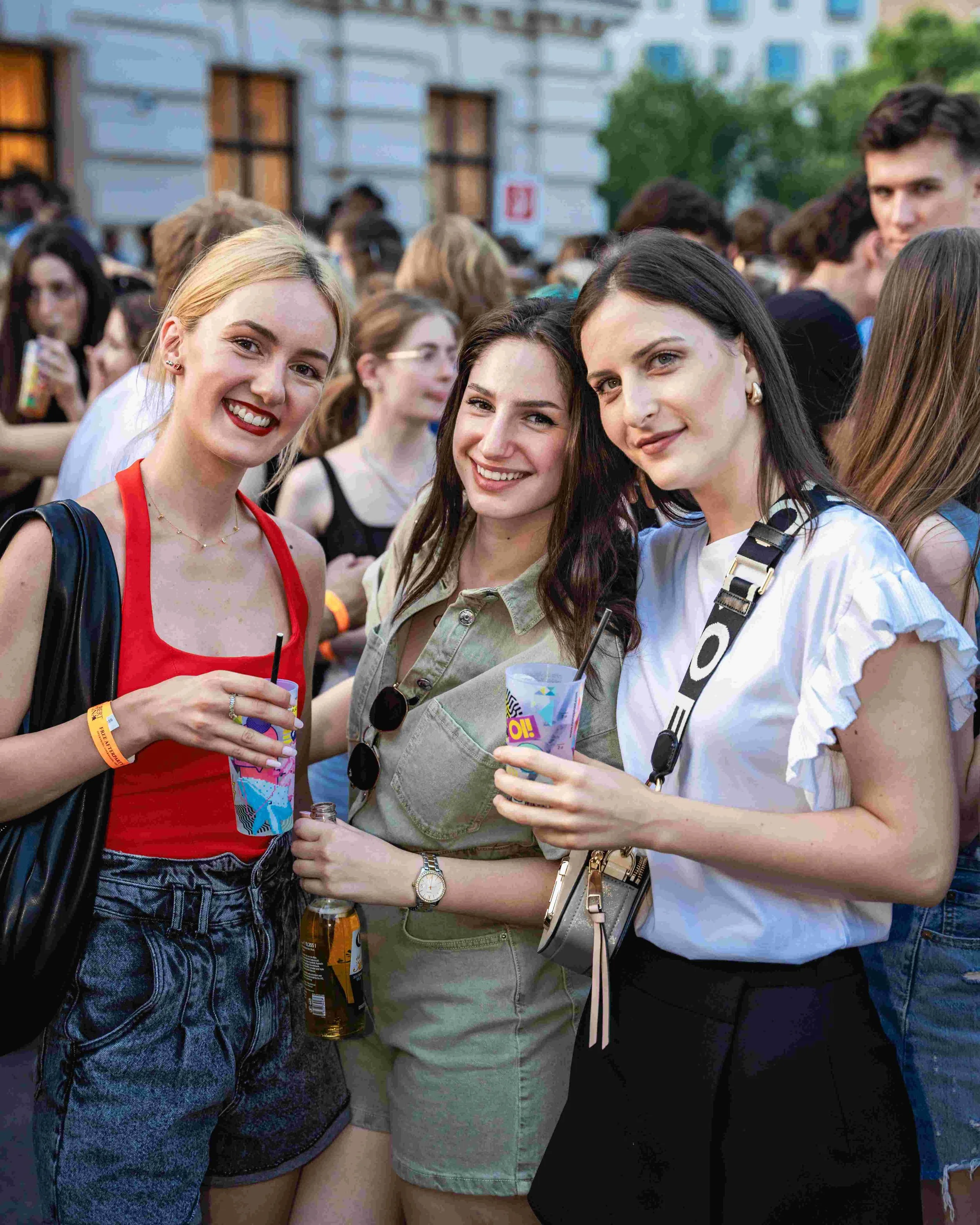 Three young women standing close together at an outdoor event, smiling and holding drinks. The woman on the left has blonde hair, a red top, and earrings. The woman in the middle has dark hair, a light-colored jacket, and sunglasses. The woman on the