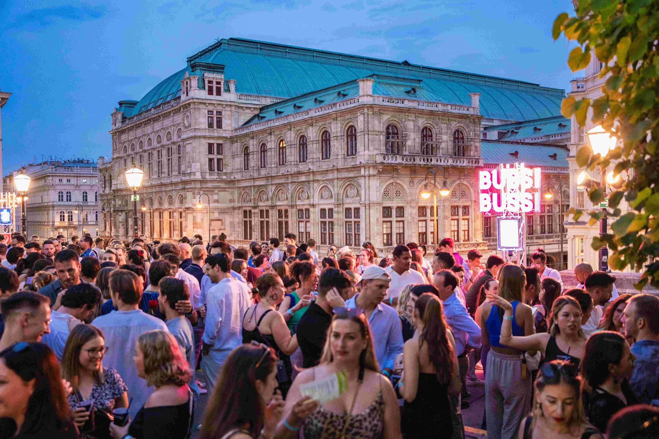 A large crowd of people gathered outdoors in front of a historic building with large windows and ornate architecture, during what appears to be evening, with some streetlights and neon signs visible.
